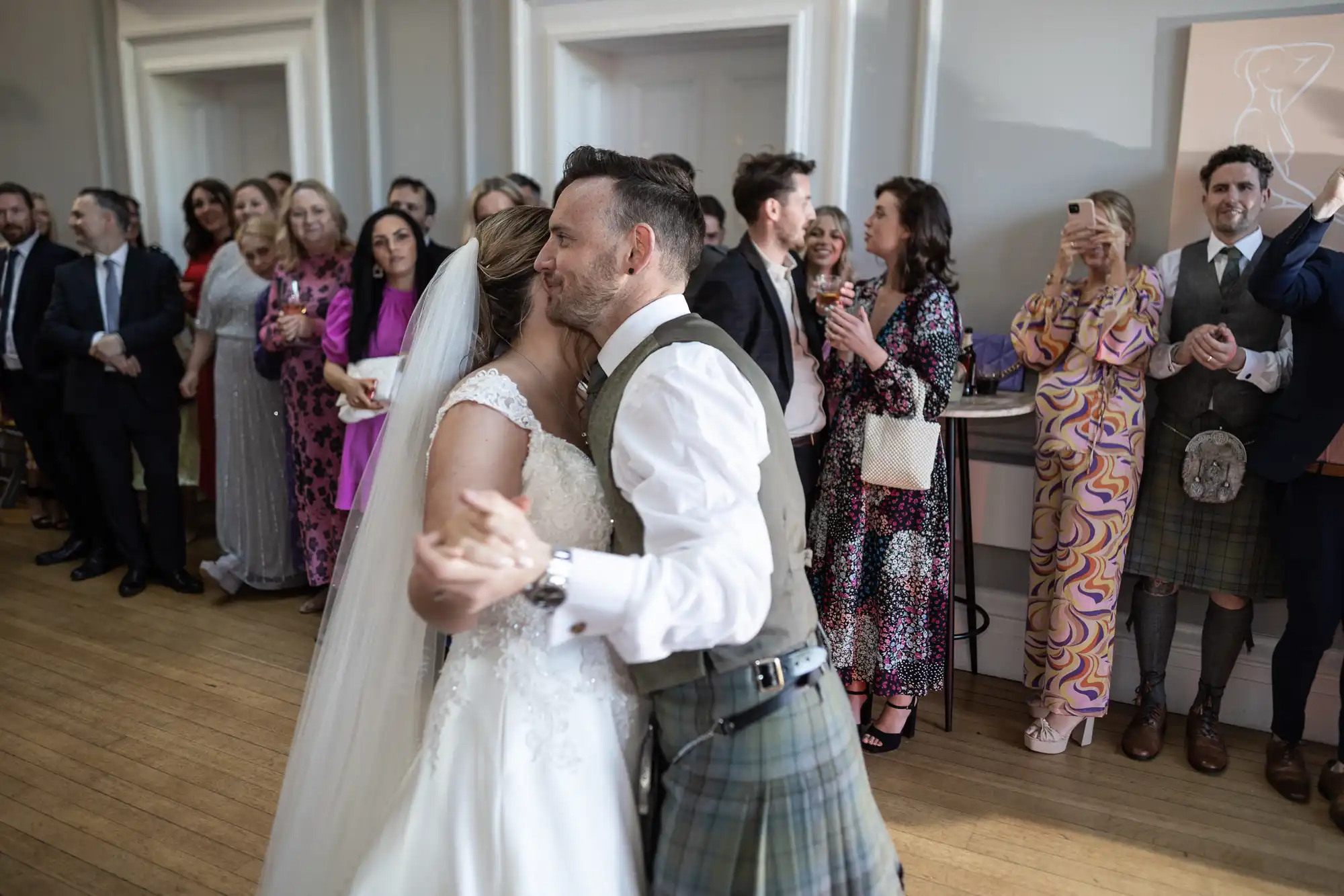 A couple in wedding attire shares a dance while guests in formal clothing observe and take photos in a well-lit room with wooden flooring.