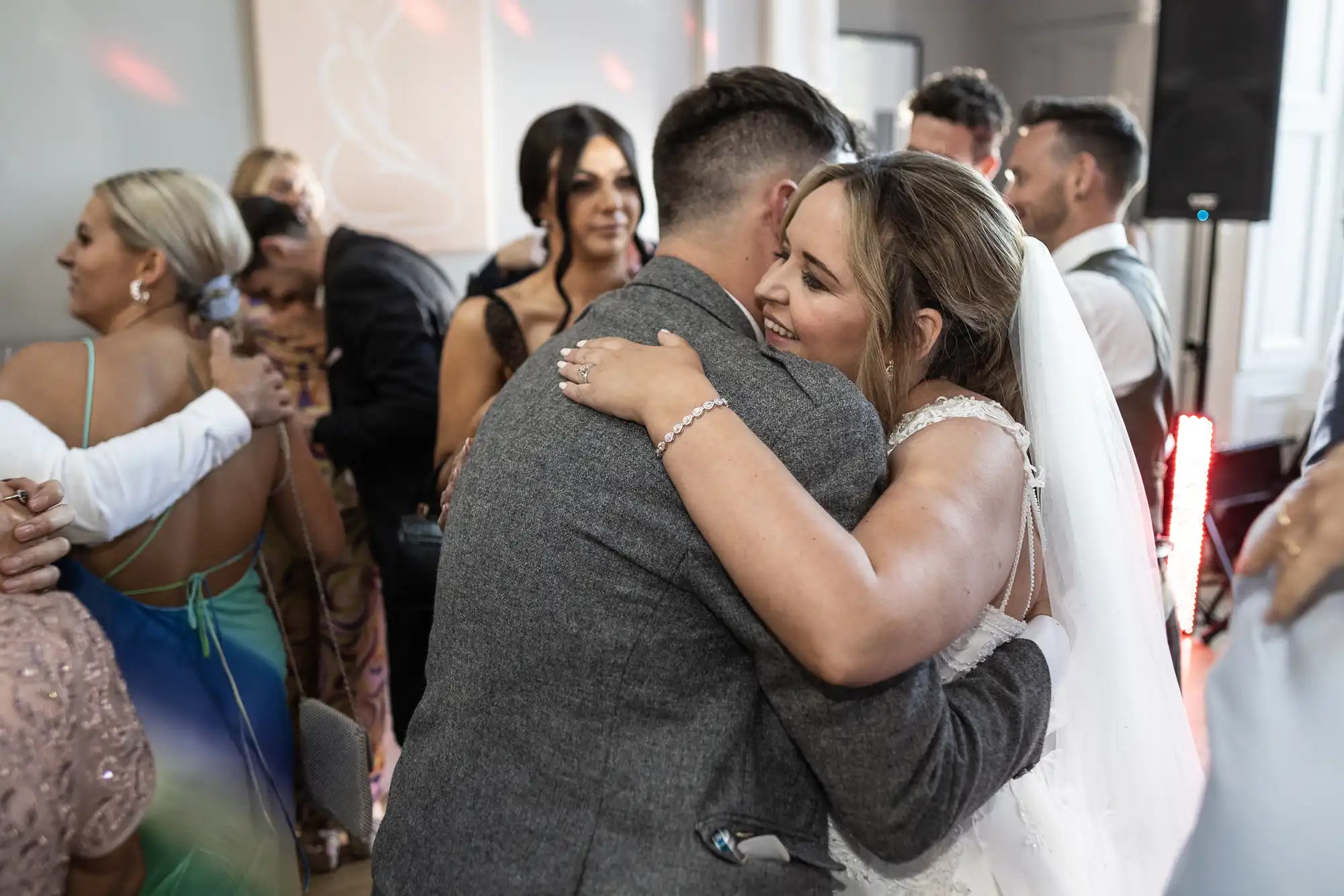 A bride in a white dress and veil hugs a man in a grey suit. Other guests in formal attire are visible in the background.