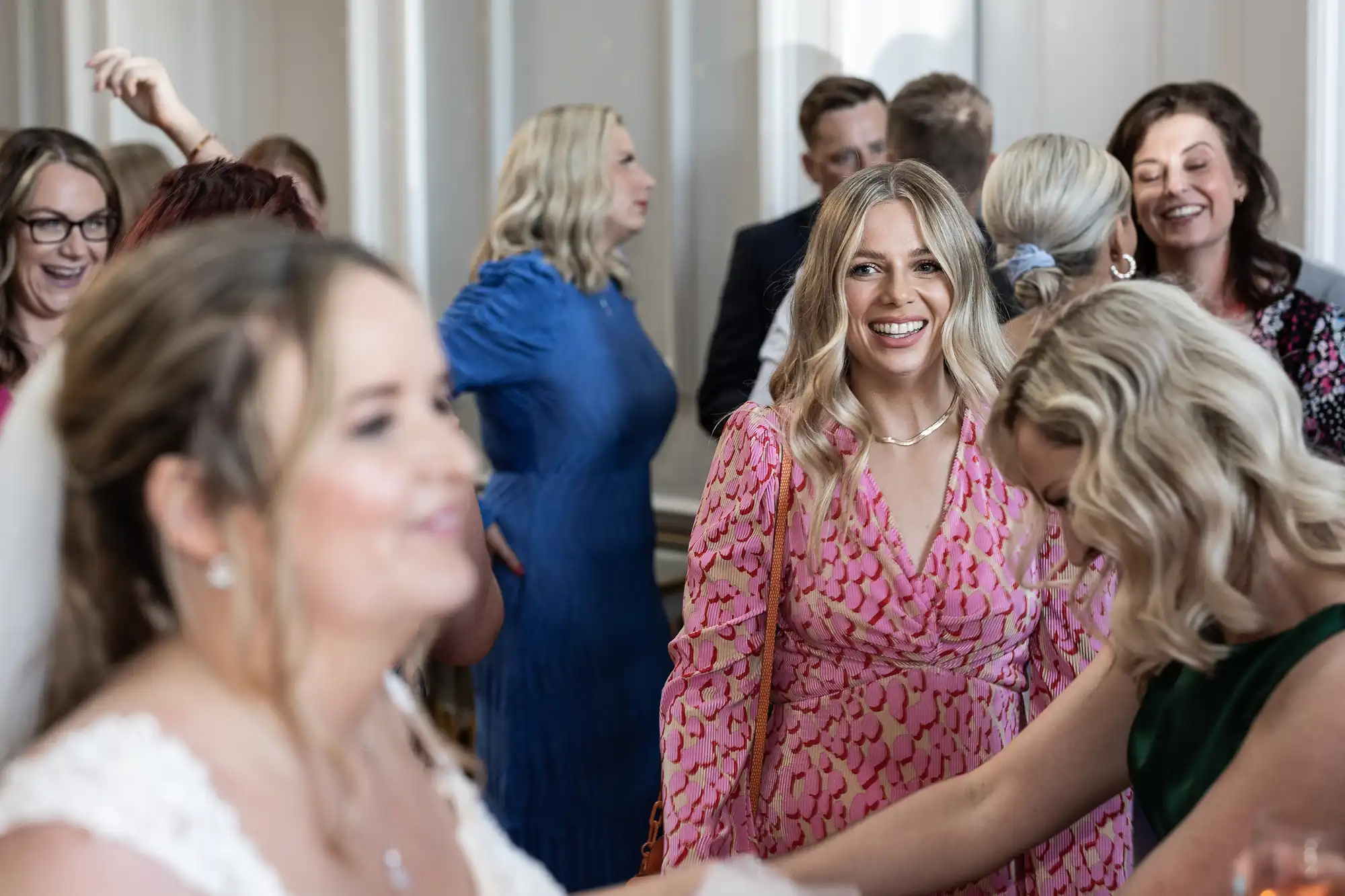 A group of people, including women in vibrant dresses, are engaged in conversation at a social gathering. The focus is on a woman in a pink dress smiling, while others converse in the background.