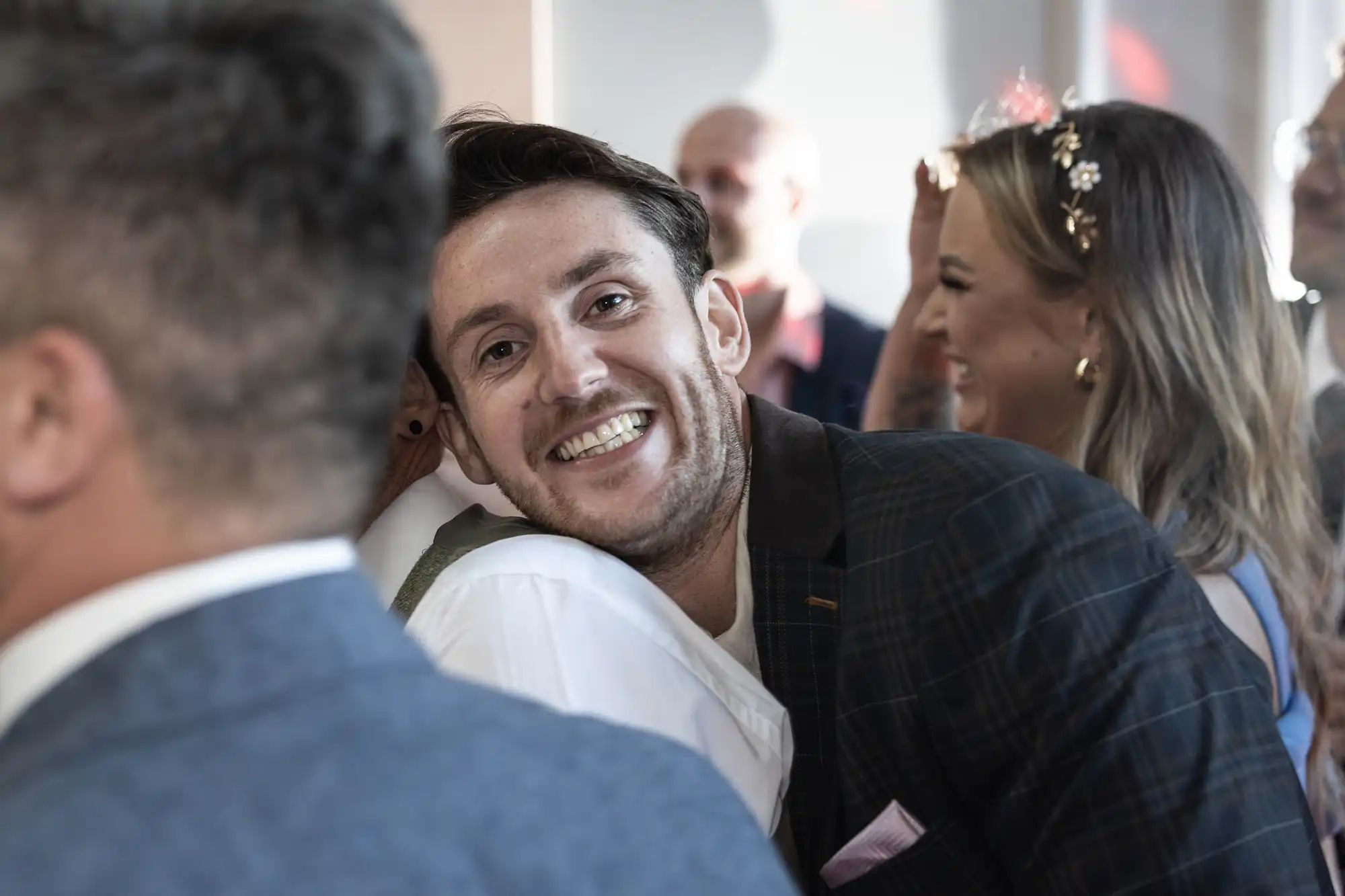 A man, smiling broadly, leans his head on someone else's shoulder in a crowded gathering. Others in the background are also engaged in conversation and laughter.