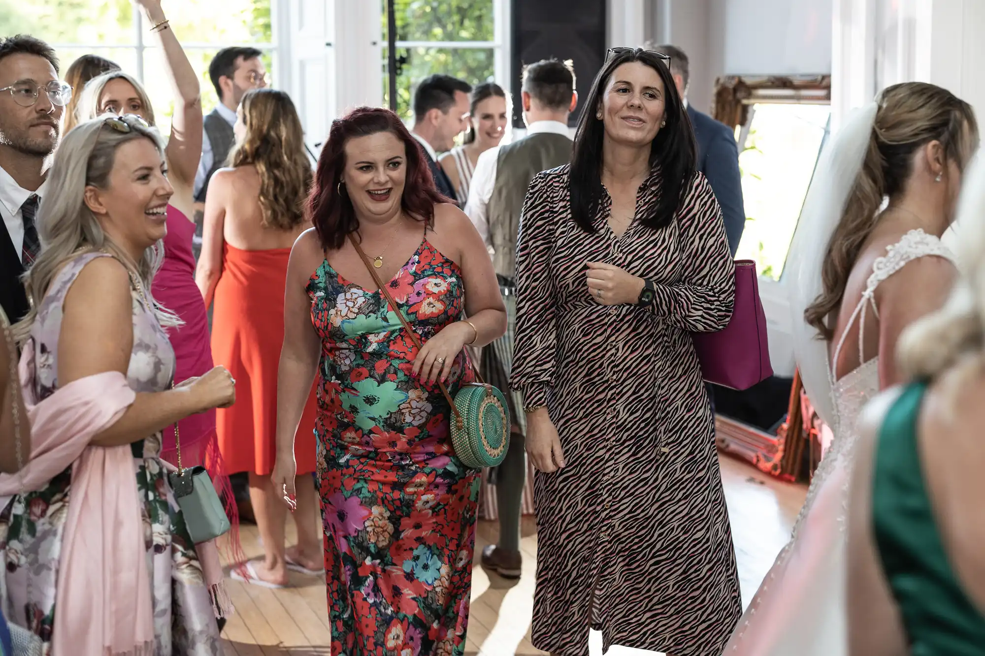 A group of people dressed in formal attire, engaging in conversation at an indoor event. One person is wearing a floral dress, and another is in an animal print outfit.