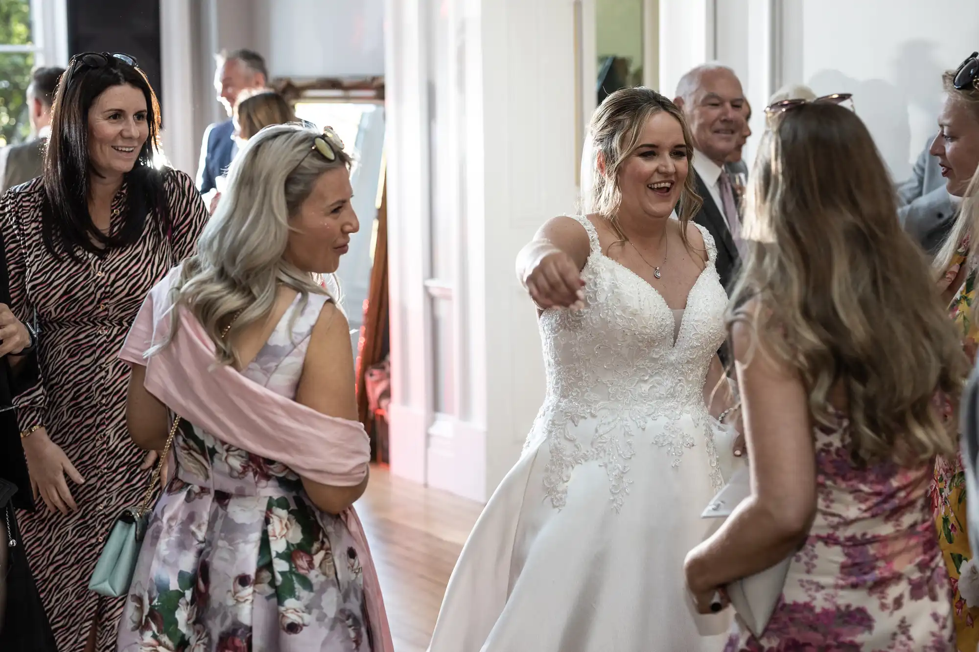 A bride in a wedding dress joyfully greets guests, including several women in floral dresses, in a bright room.