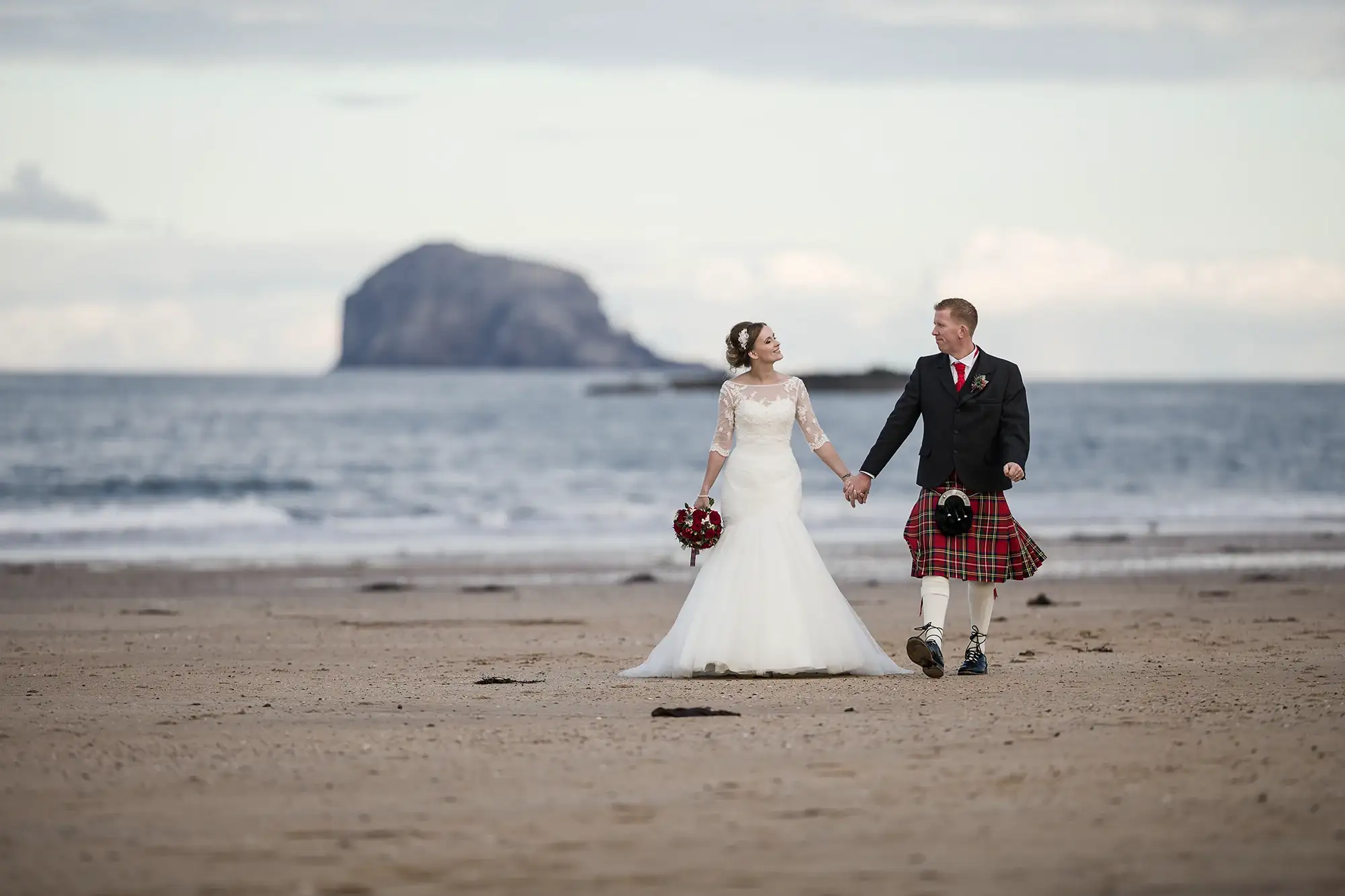 A bride in a white dress and a groom in a kilt hold hands on a beach, with a large rock formation in the distant sea behind them.