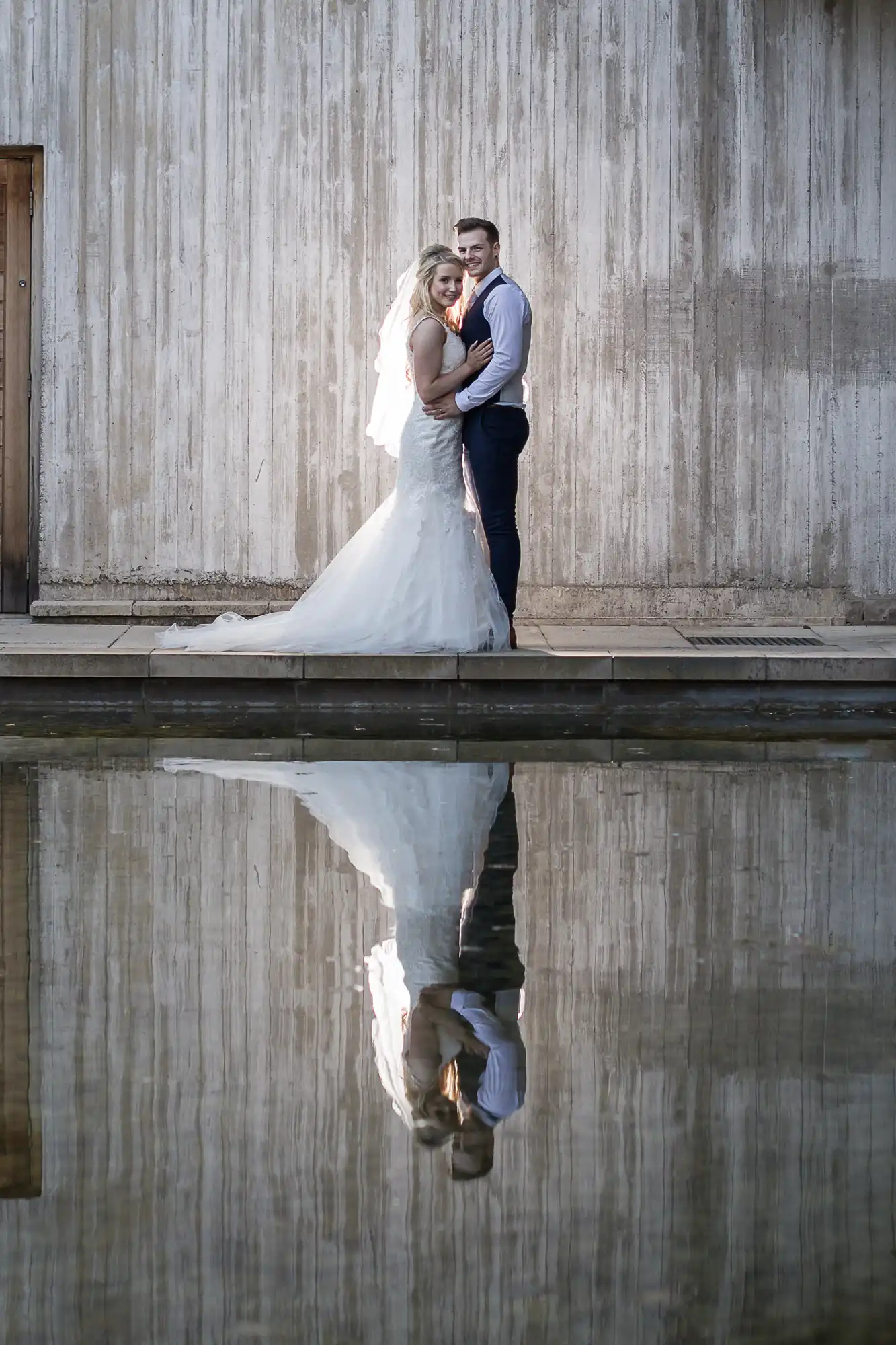 A bride and groom stand by a concrete wall, reflected in a still body of water. The bride wears a white gown; the groom is in a light shirt and dark pants.