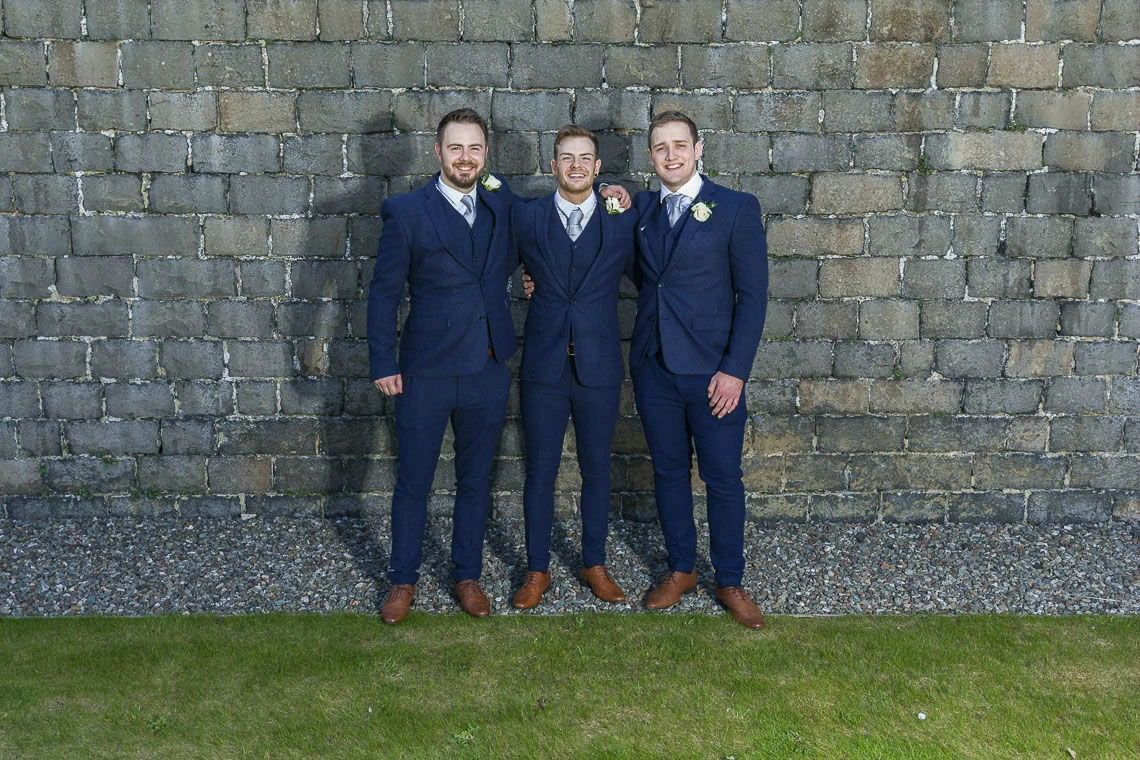 Groom and bestmen pose for photo against stone wall in garden