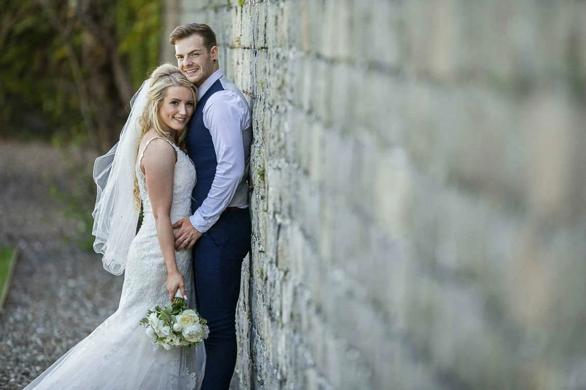 bride and groom pose against stone wall