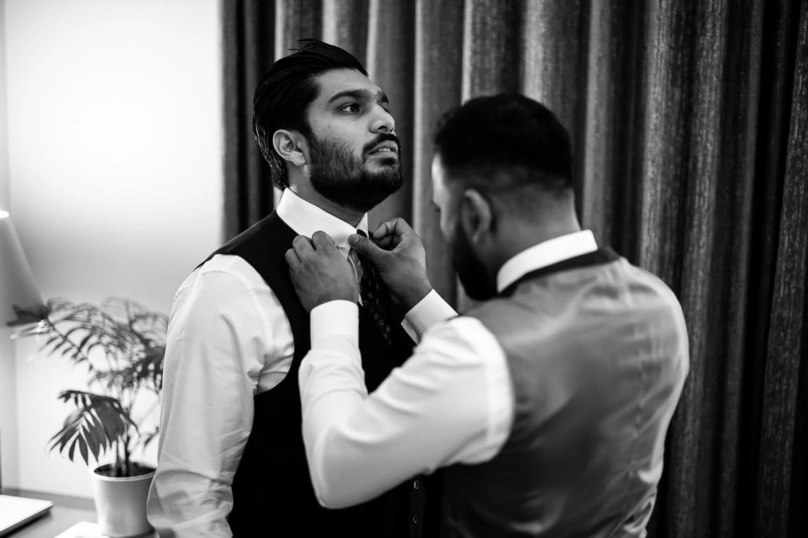 groomsman helps groom put on a tie
