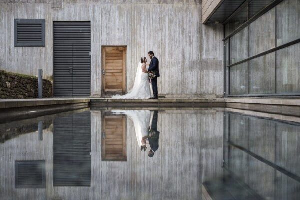 Norton House Hotel wedding photography - relaxed and natural couple portraits by Norton House Hotel wedding photographer: A bride and groom stand close together by a wooden door, their reflection visible in a calm, shallow pool in front of a modern building.