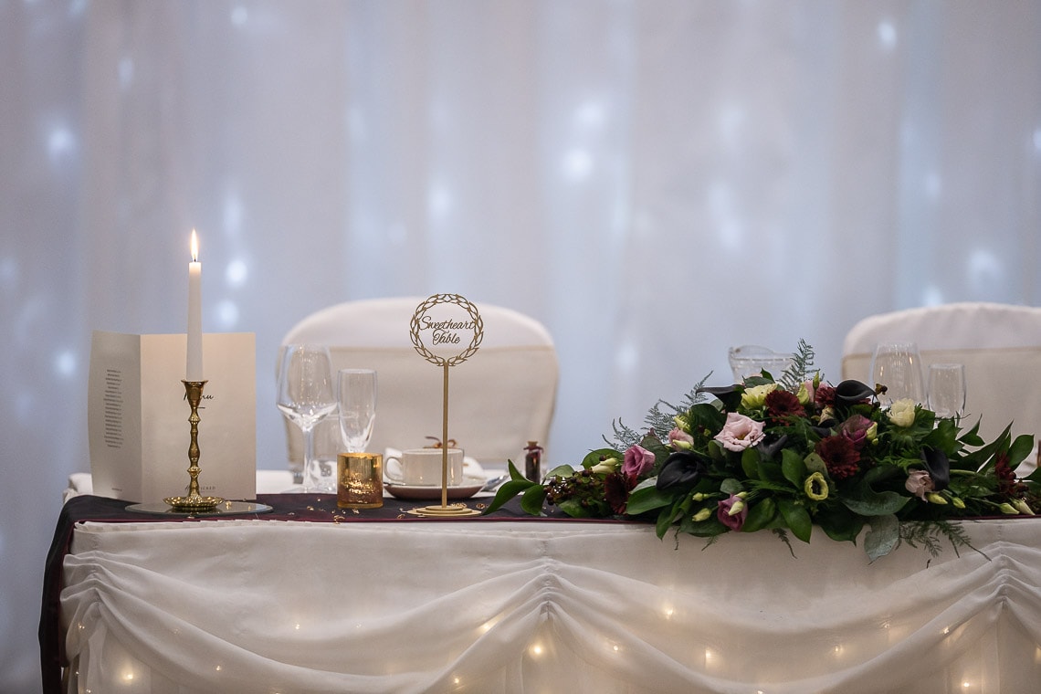 Elegant wedding table setting with a floral centerpiece, candlestick, two glasses, and a place card reading "splendid bride" against a draped, fairy-lit backdrop.