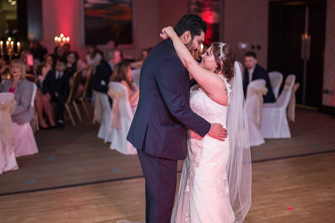 A newlywed couple sharing their first dance in a ballroom surrounded by seated guests watching them.
