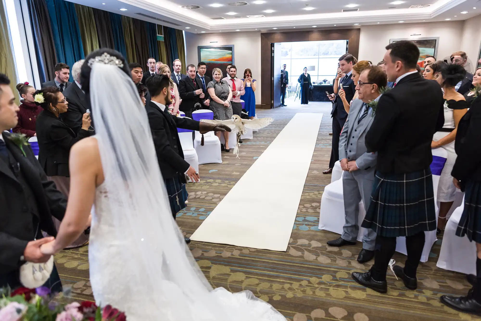 A bride and groom stand at the end of an aisle inside a decorated room, surrounded by guests watching an owl fly towards them.