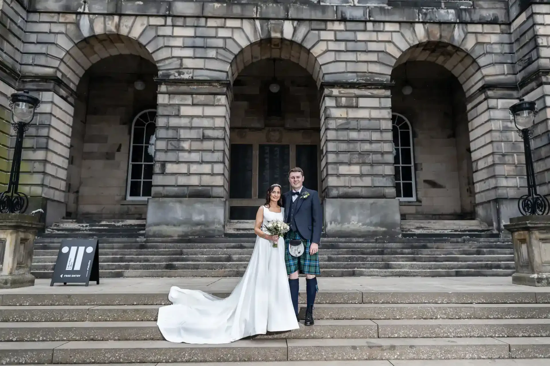 A bride in a white gown and a groom in a kilt pose together on stone steps in front of a historic building with large arches and stone columns.