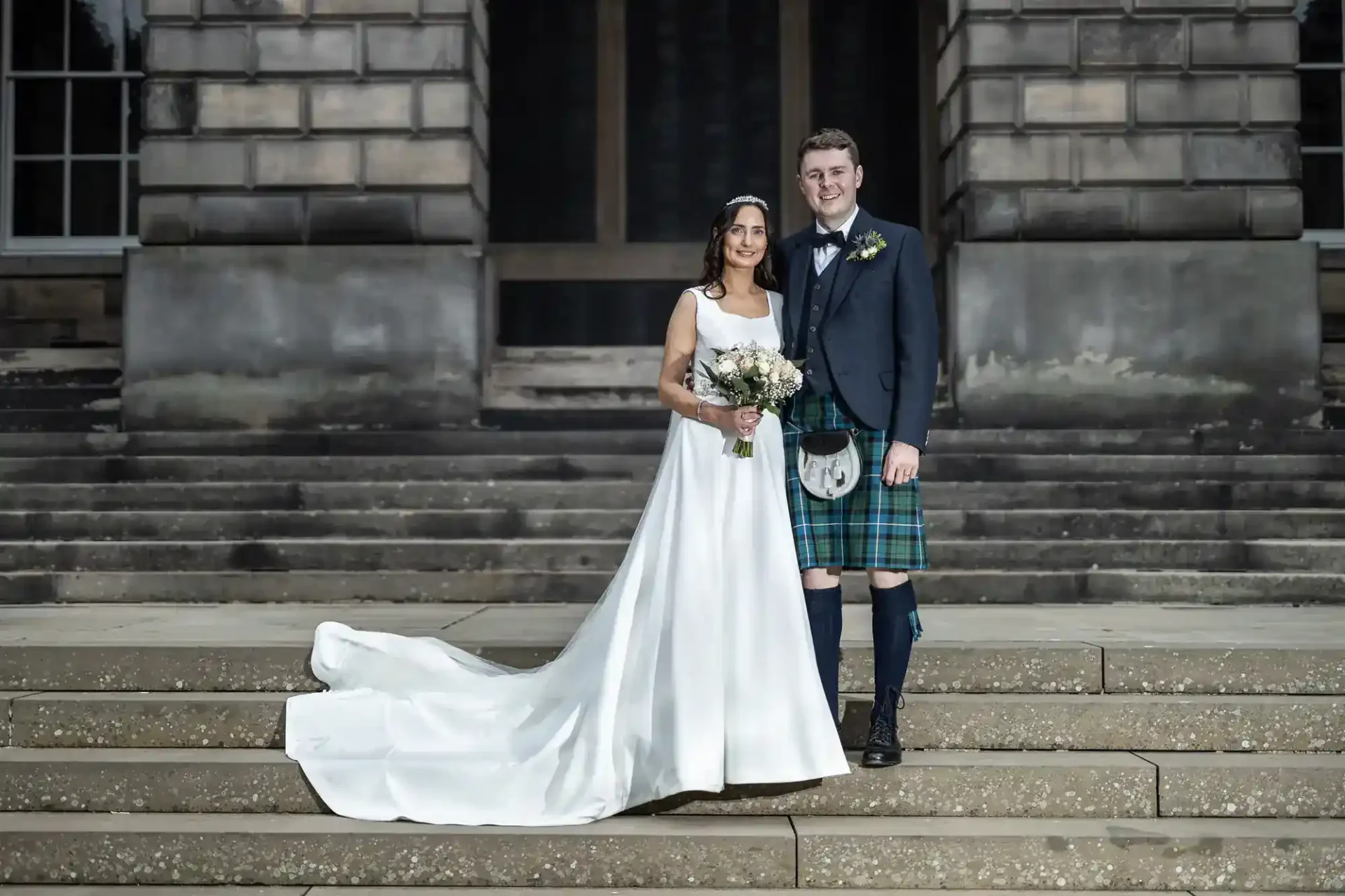 A bride in a white gown and a groom in a kilt stand side by side on stone steps in front of a large building, posing for a wedding photo.