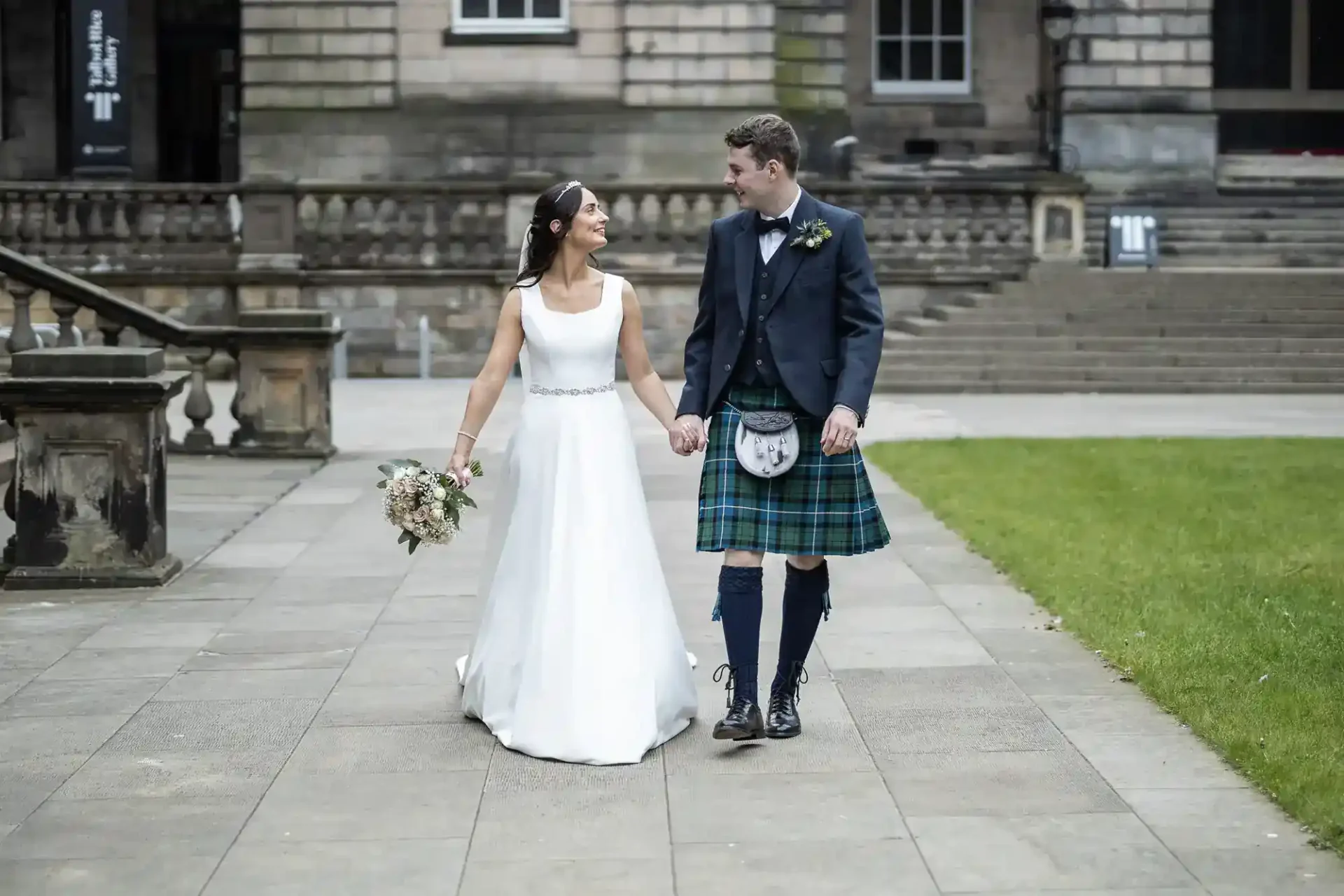 A bride in a white dress and a groom in a kilt and jacket walk hand in hand outside a stone building, looking at each other.