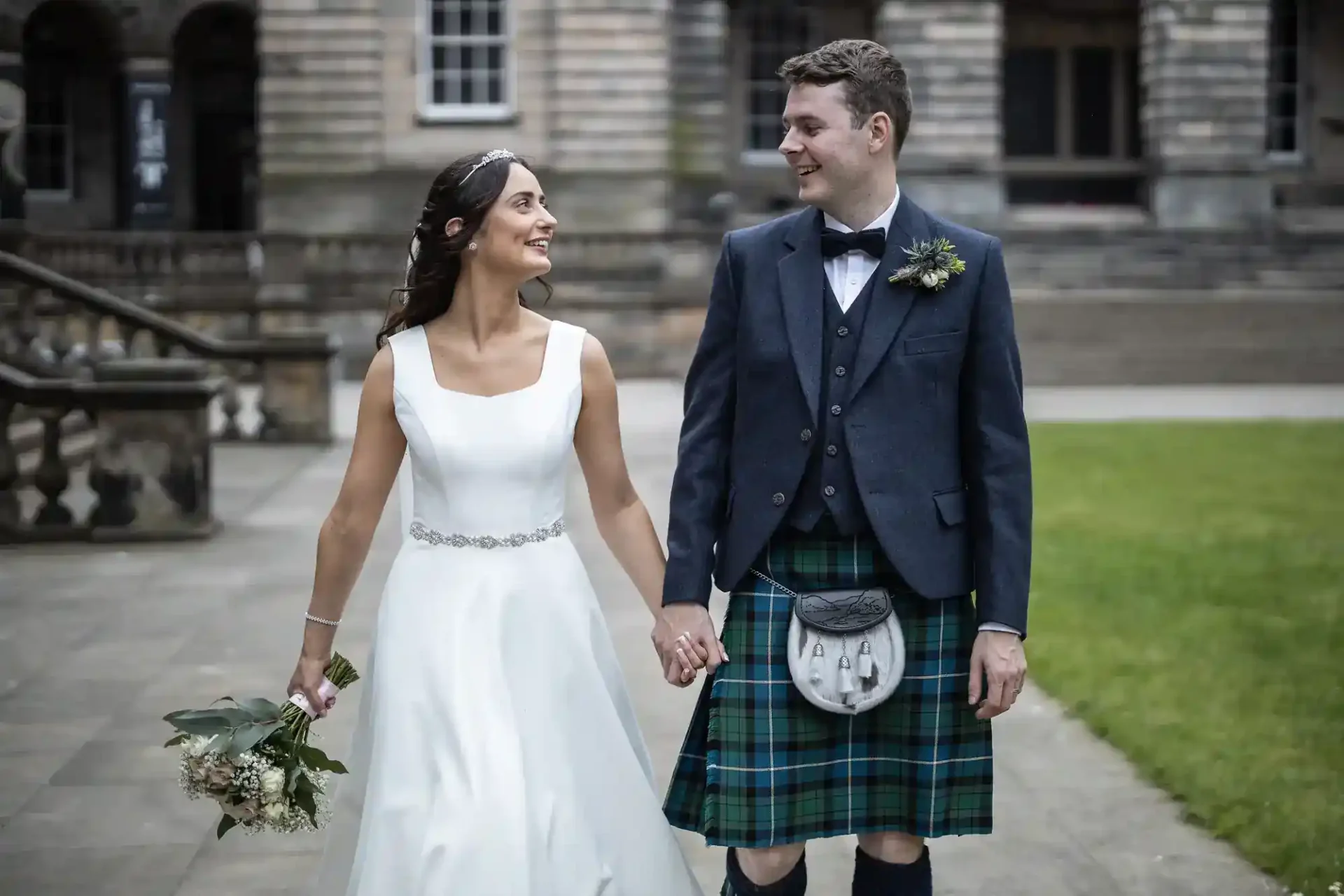 A bride in a white dress and a groom in a kilt hold hands and walk outside a historic stone building, smiling at each other.