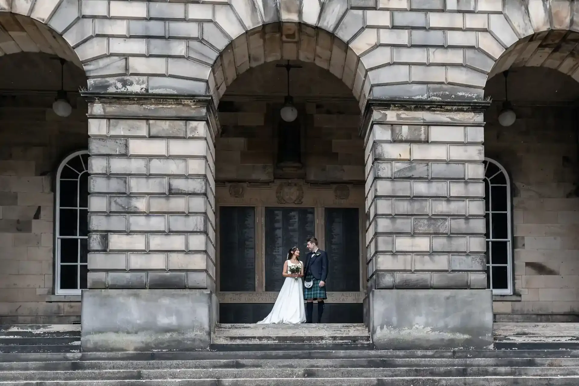 A bride and groom stand together under a large stone archway, posing for a wedding photo in front of a historic building.