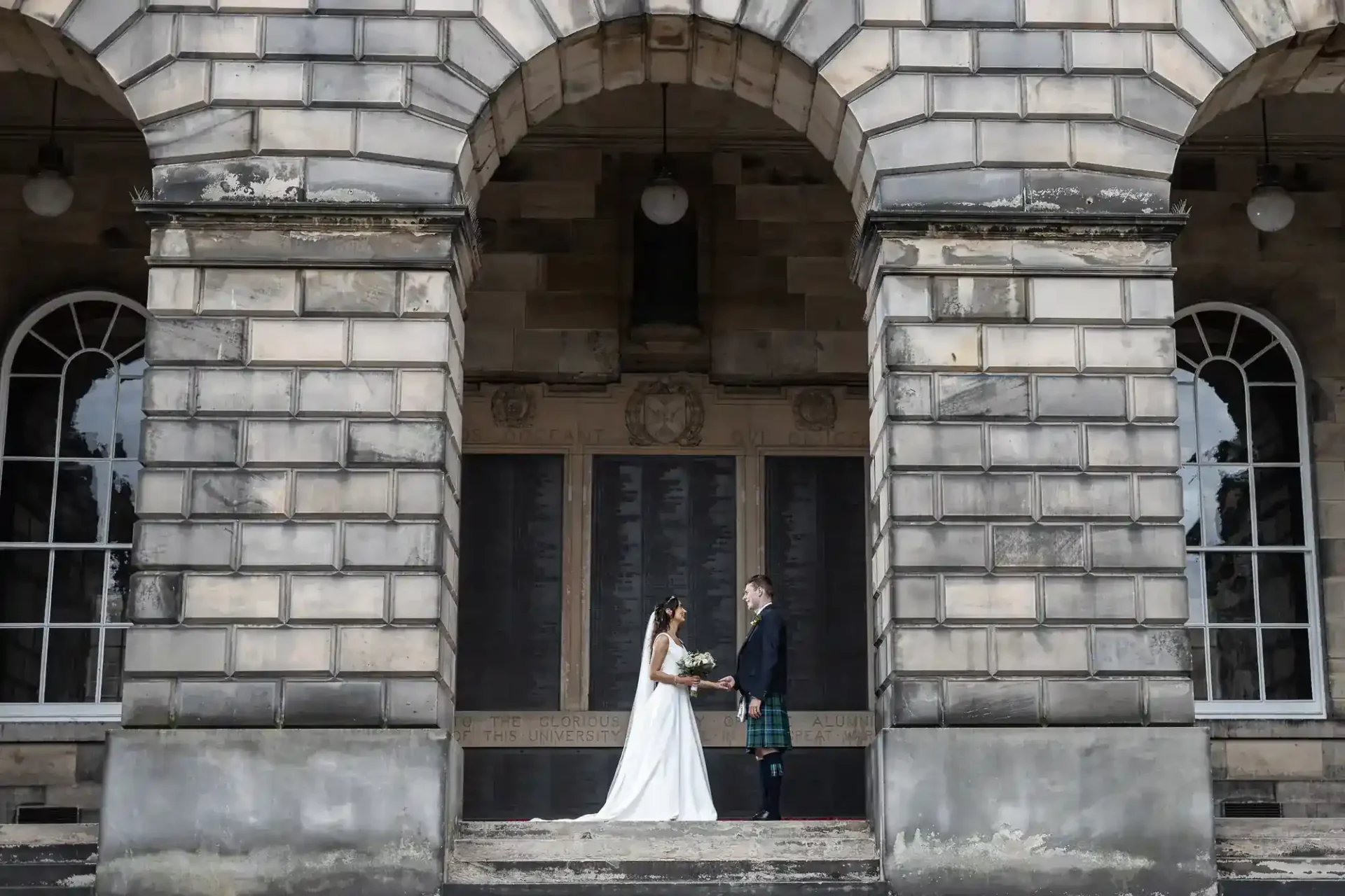 A bride and groom stand facing each other under a large stone archway, holding hands, with the building’s stone facade visible behind them.