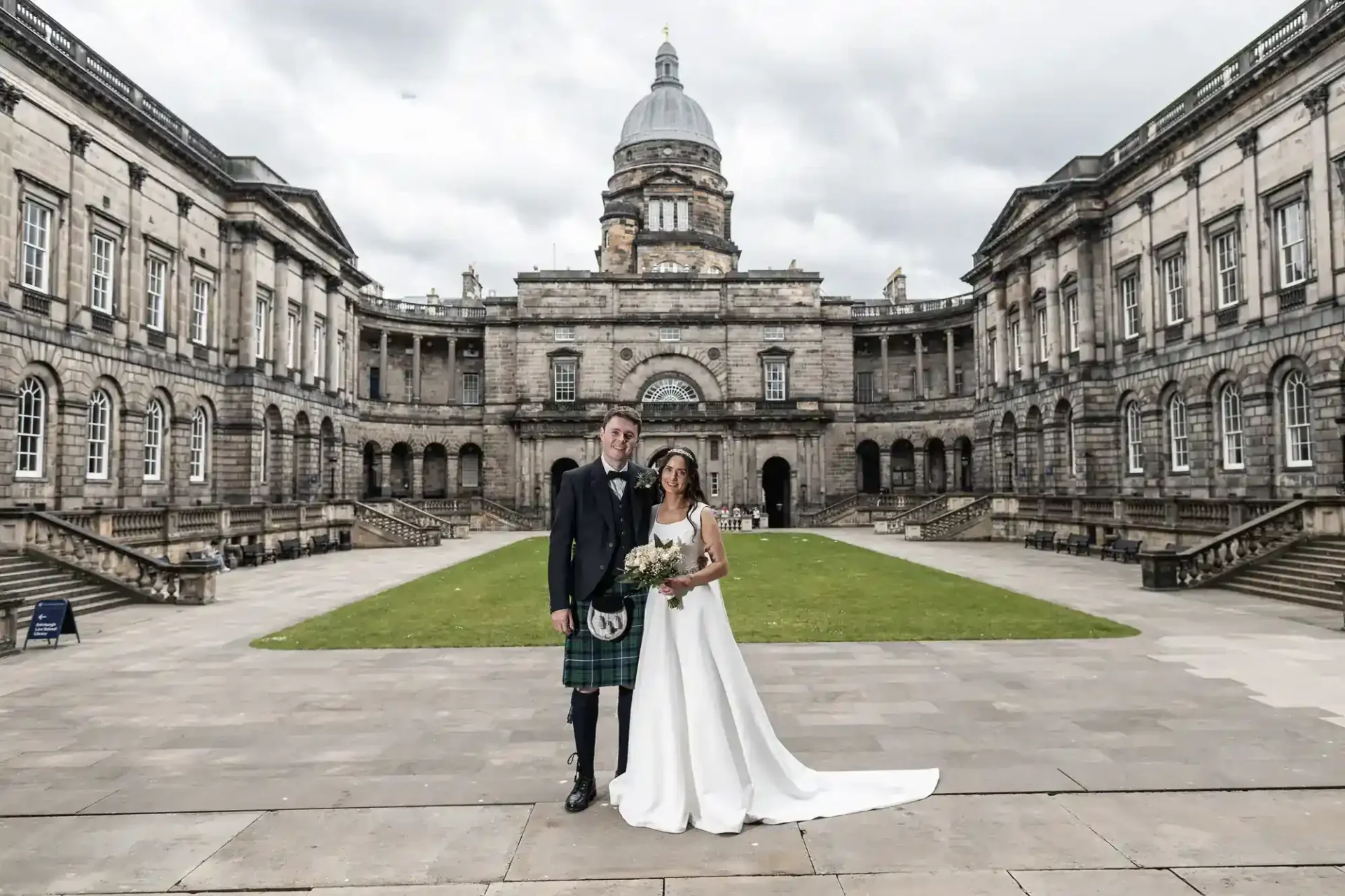 A bride and groom pose for a wedding photo in the courtyard of a grand historic building with a domed tower and stone architecture.