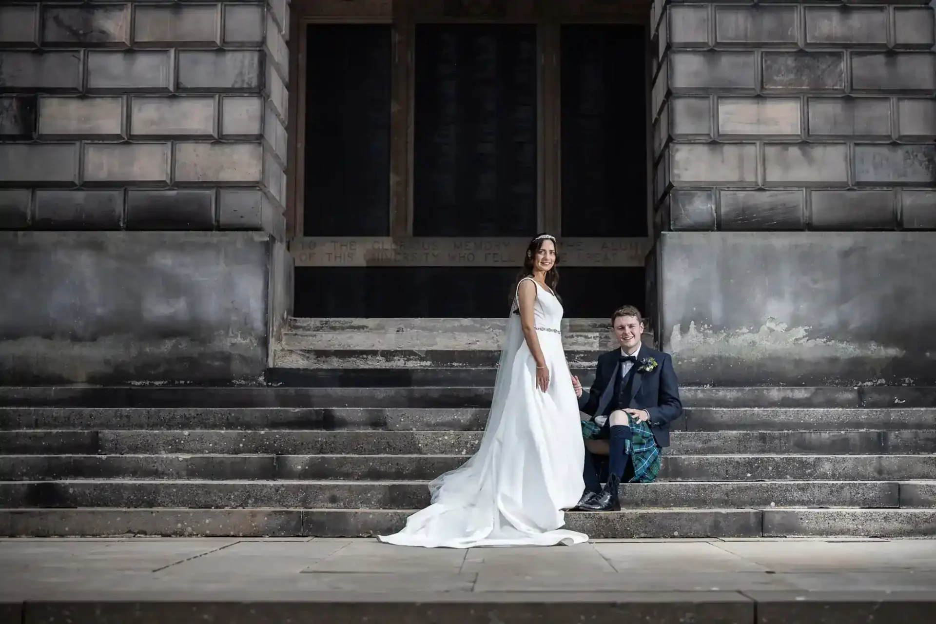 A bride in a white gown stands beside a groom in a suit and kilt, seated on stone steps in front of a large stone building.
