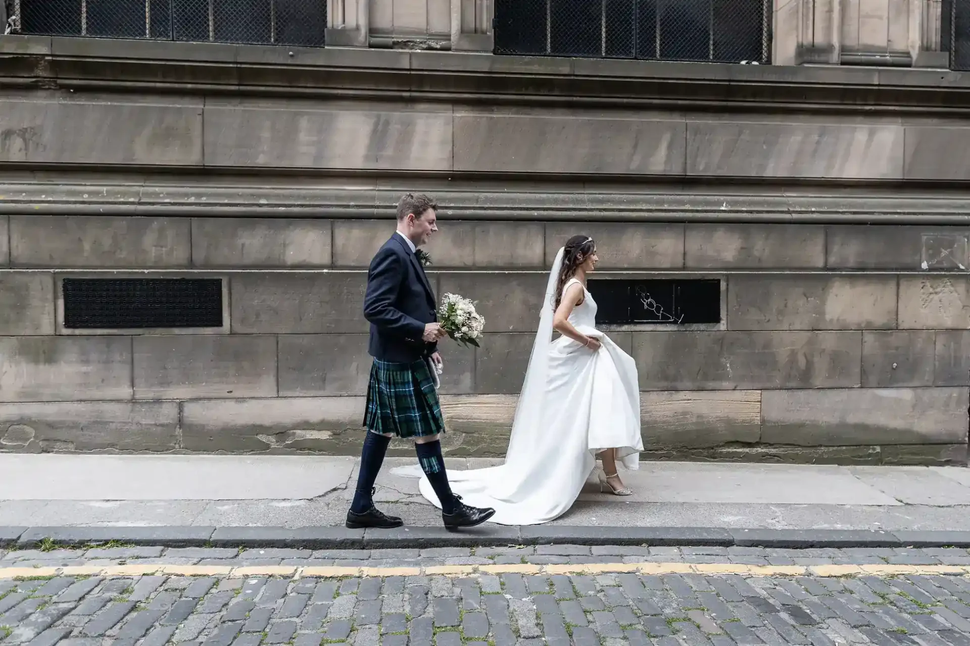 A bride in a white dress and a groom in a kilt walk together along a stone sidewalk beside a historic building.