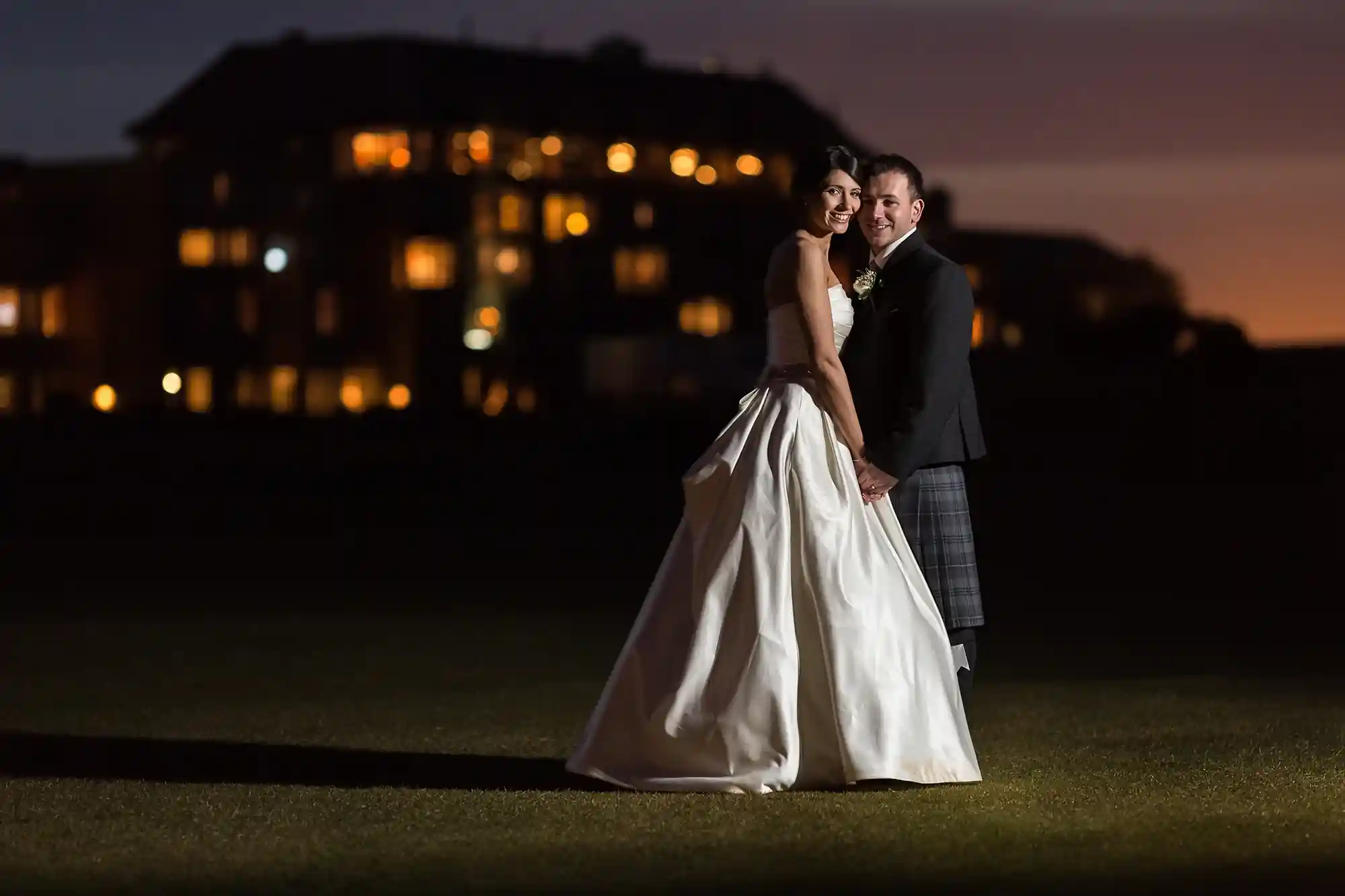 A newlywed couple, the bride in a white gown and the groom in a kilt, posing at dusk with a dimly lit building in the background.