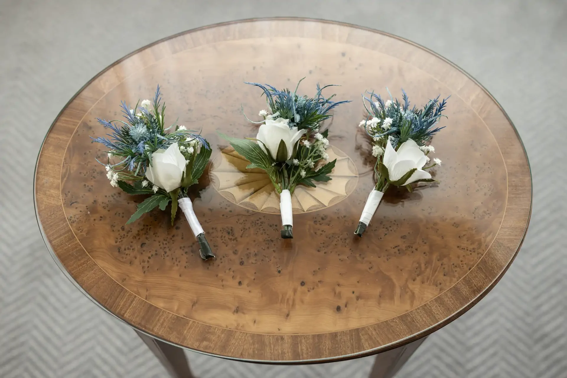 Three boutonnières with white roses, blue thistles, and greenery are arranged on a round wooden table.