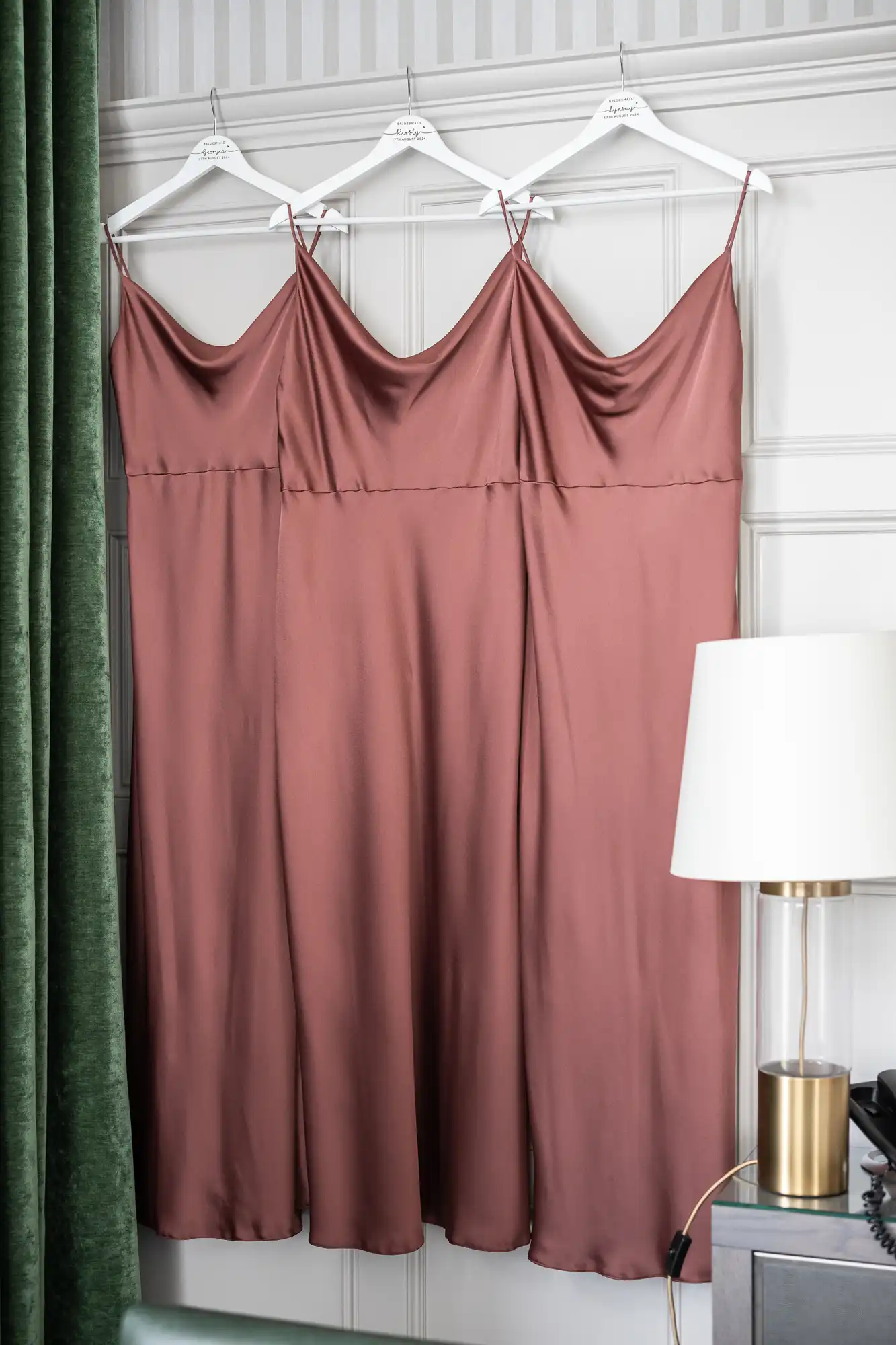 Three identical mauve satin slip dresses hang on white hangers against a paneled wall near a lamp and green curtain.