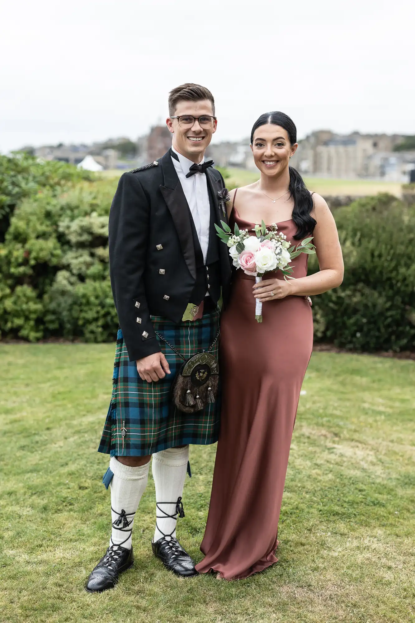 A man in a kilt and a woman in a long dress pose together outdoors on grass, with buildings in the background.