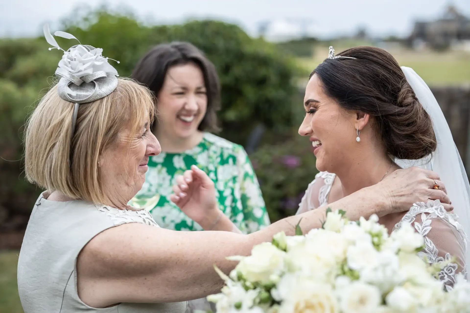 Bride smiling as a woman adjusts her veil, with another woman laughing in the background.