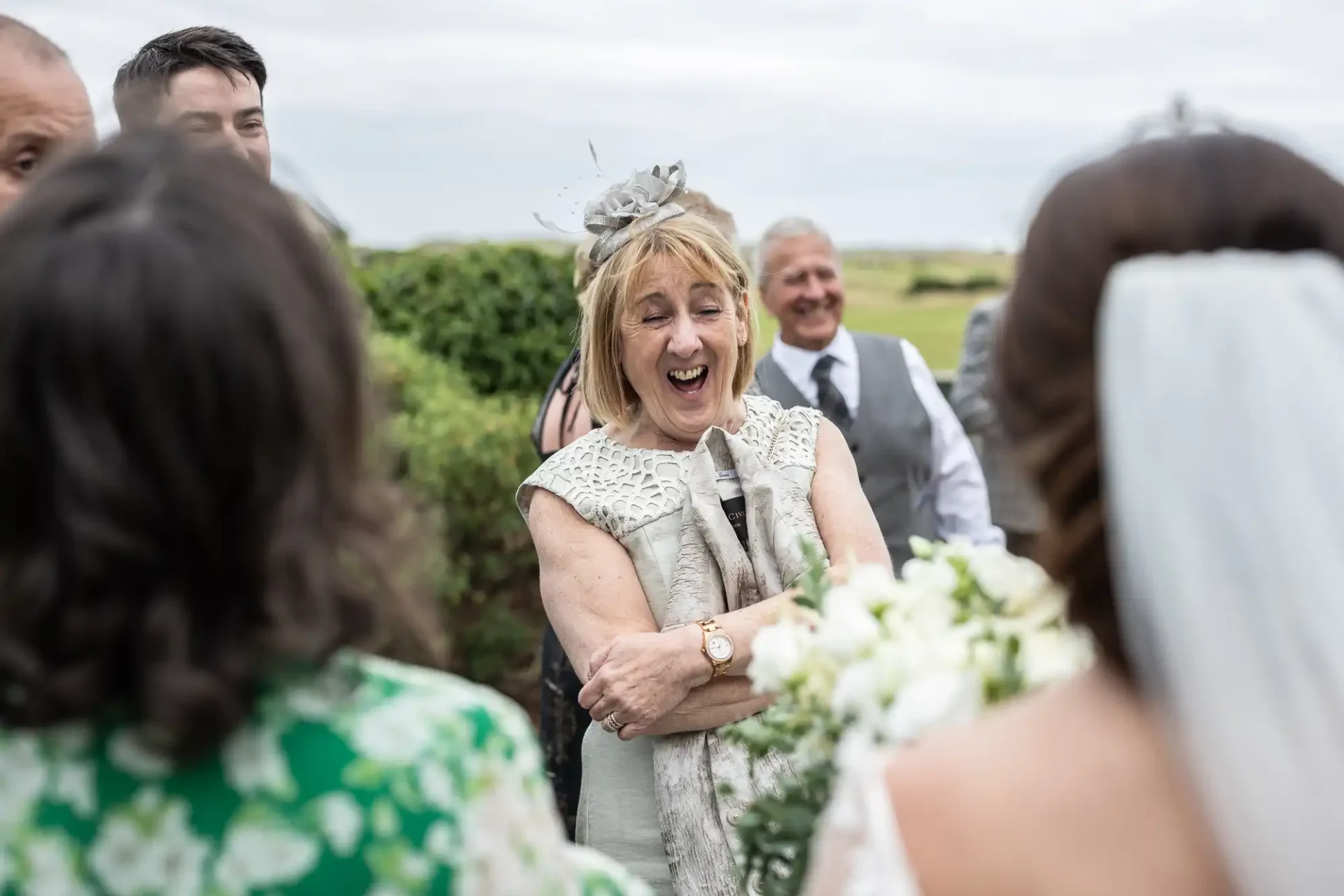 A woman with a fascinator smiles widely at an outdoor gathering, surrounded by people.