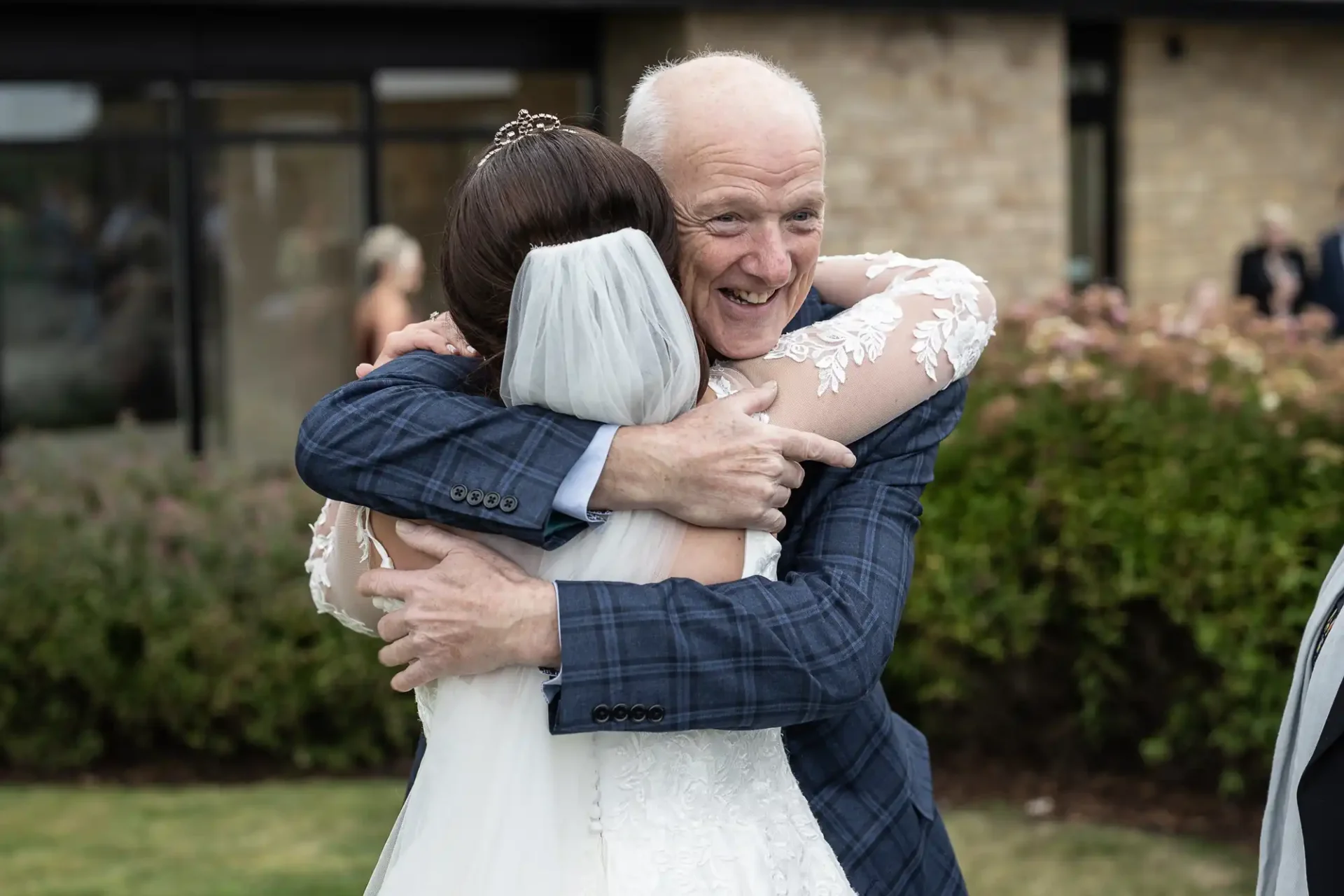 A bride in a white gown and veil hugs an older man in a blue checkered suit outside a building.