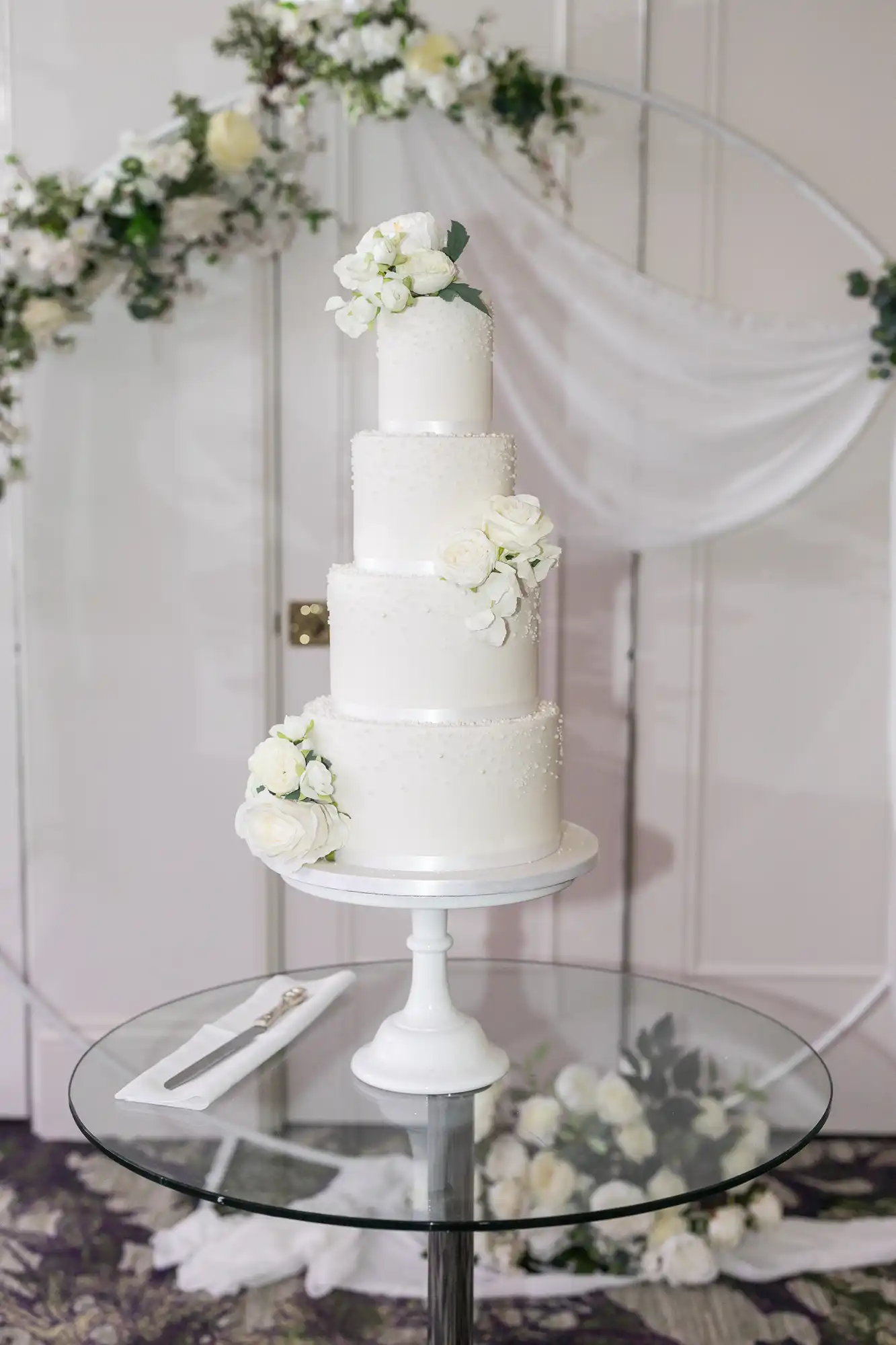 Three-tier white wedding cake with white floral decorations on a white stand, placed on a glass table. A knife and fork are beside the cake. A circular floral arch is in the background.