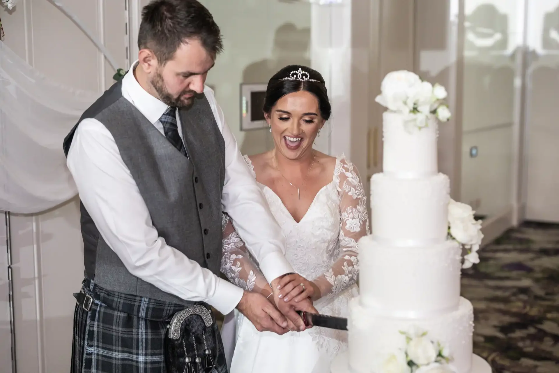 A bride and groom are cutting a tiered white wedding cake together. The groom wears a kilt, and the bride is in a lace wedding dress and tiara.