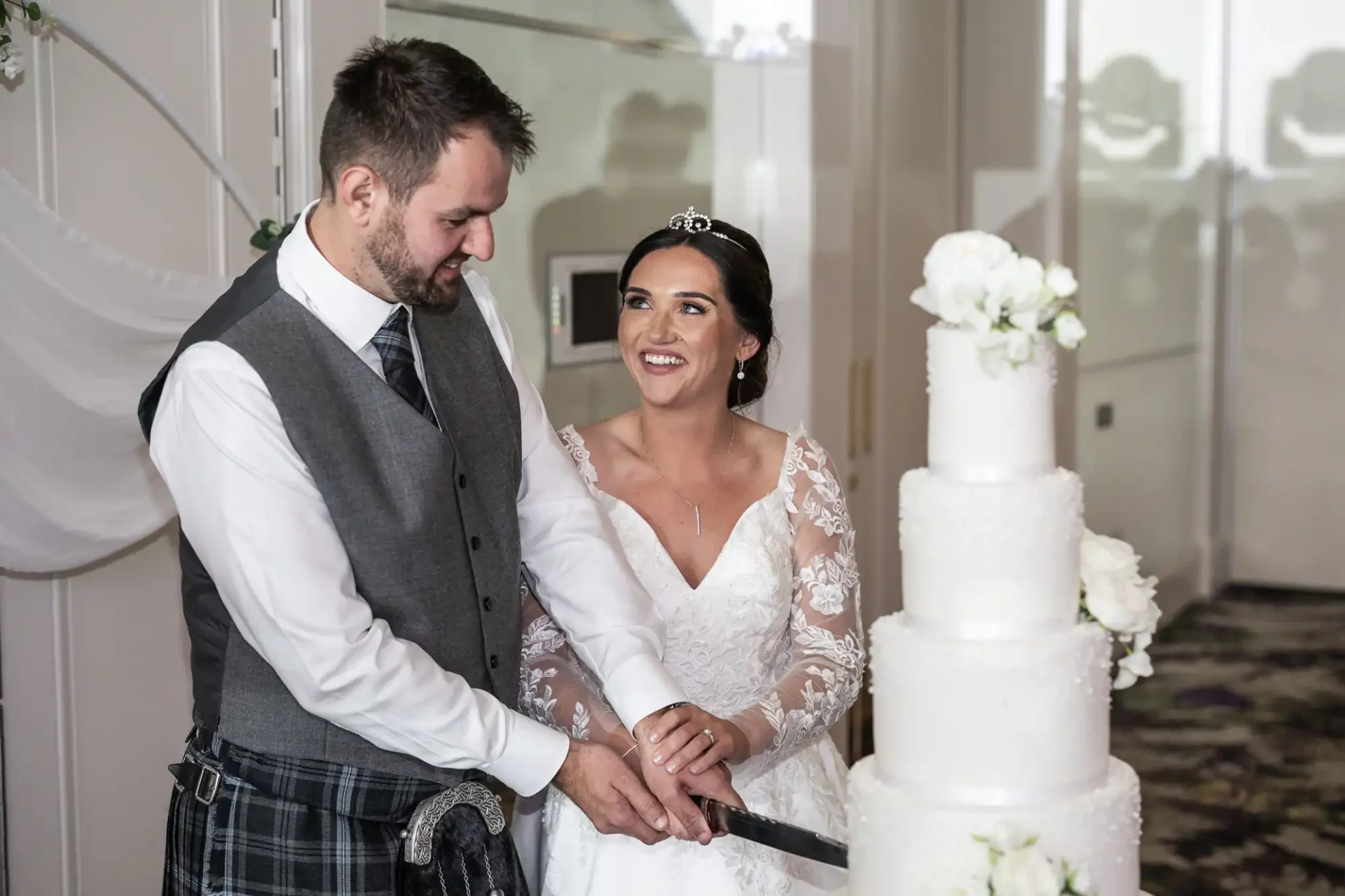 A bride and groom smile while cutting a tiered wedding cake adorned with white flowers. The groom wears a vest and kilt, and the bride wears a tiara and lace dress.