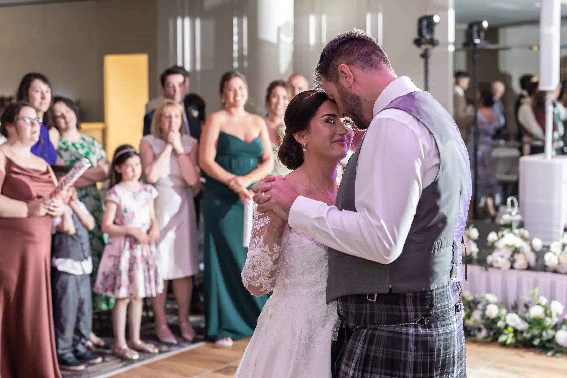 A bride and groom share a first dance surrounded by guests in formal attire.