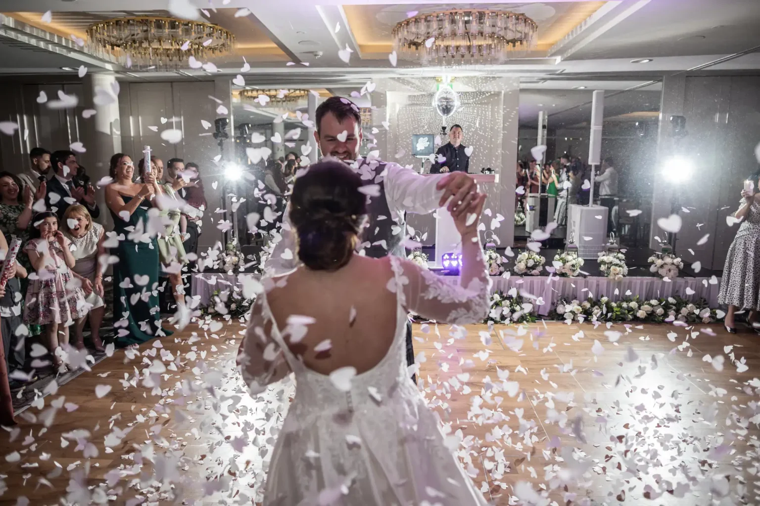 A bride and groom dance surrounded by falling white confetti on a wooden floor, while guests watch and take photos in a decorated reception hall.