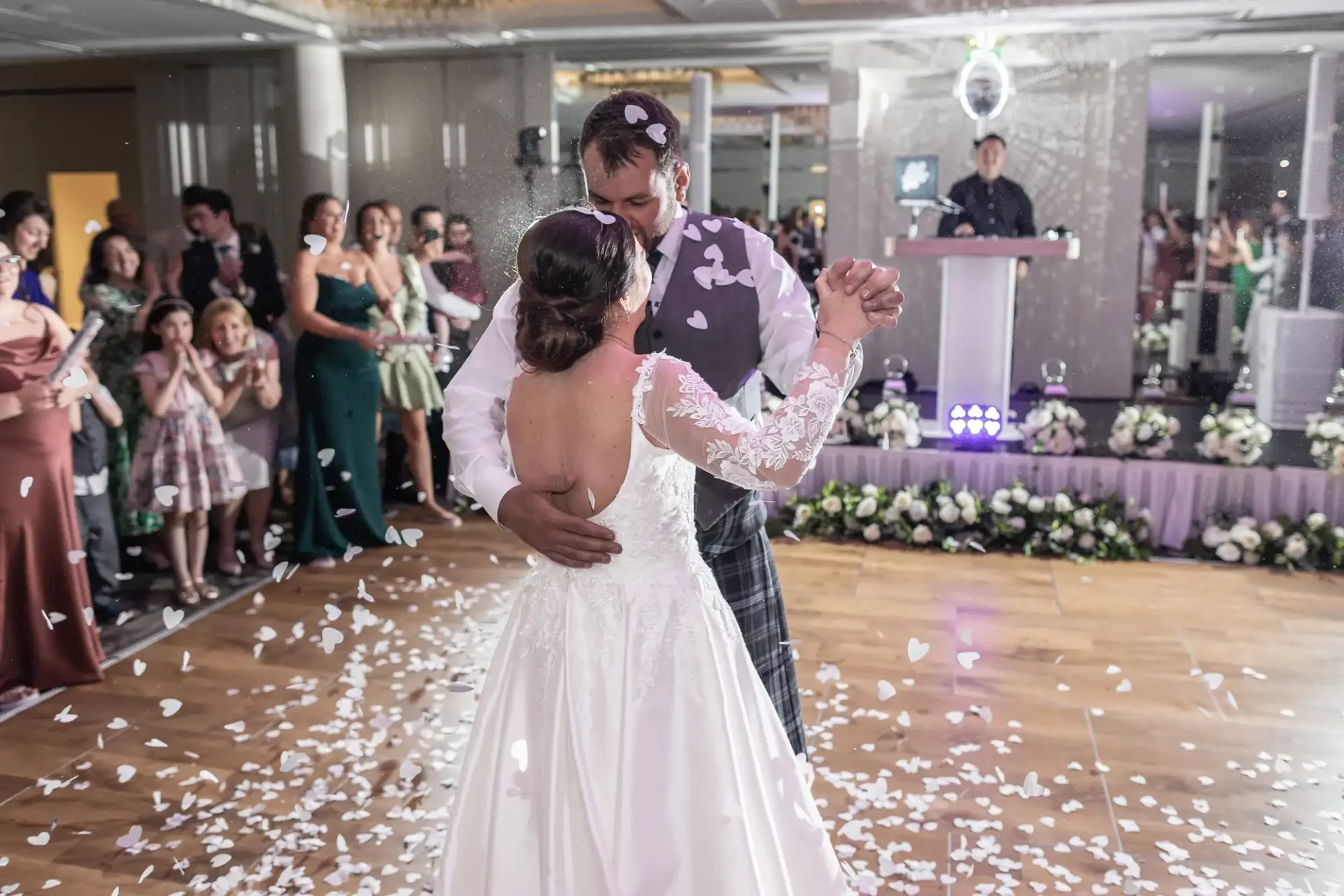 A bride and groom share their first dance on a confetti-covered floor at a wedding reception. Guests watch and a DJ stands in the background.