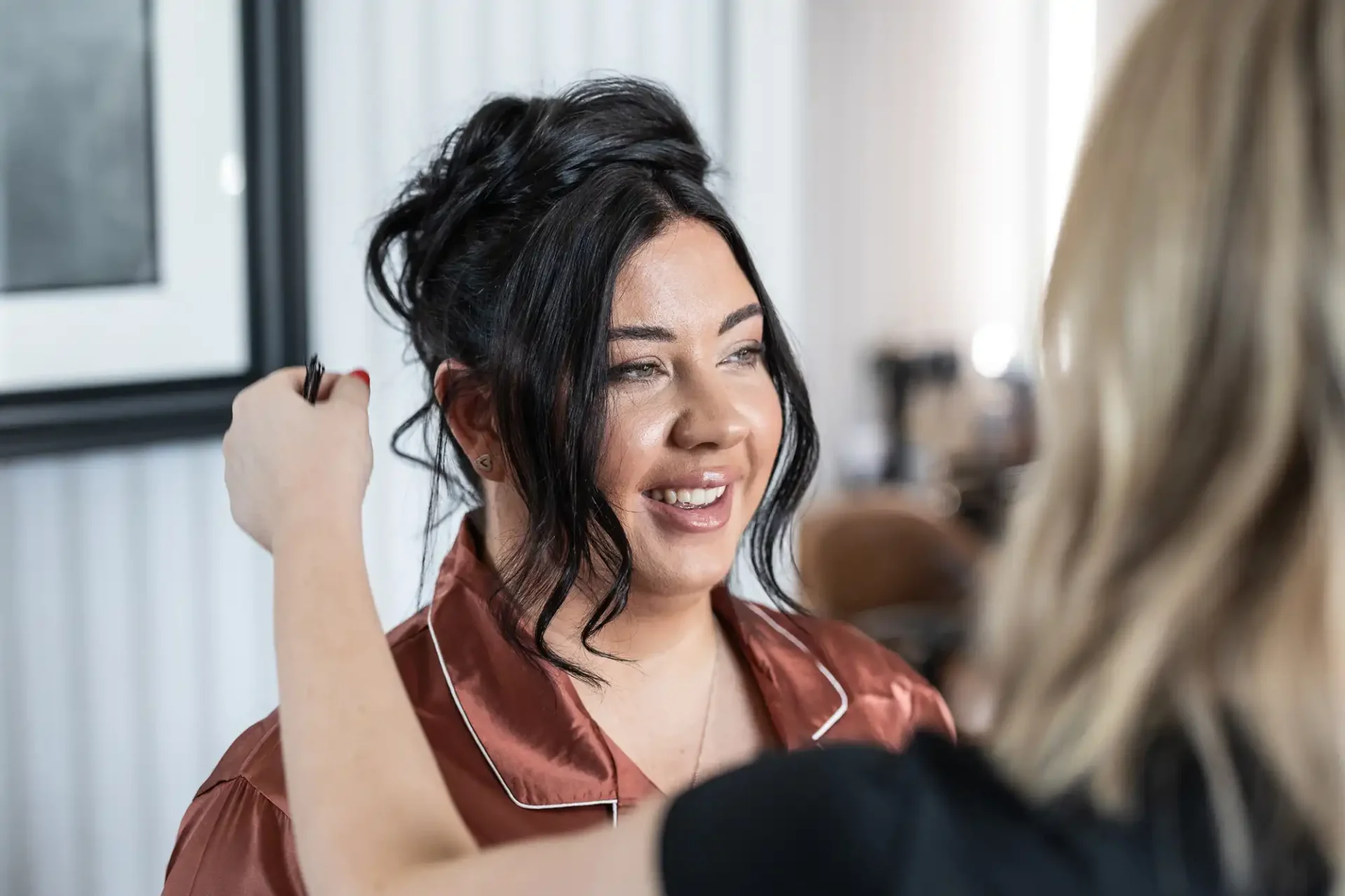 A woman with dark hair styled up is smiling as another person, with blonde hair, adjusts her appearance using makeup tools.