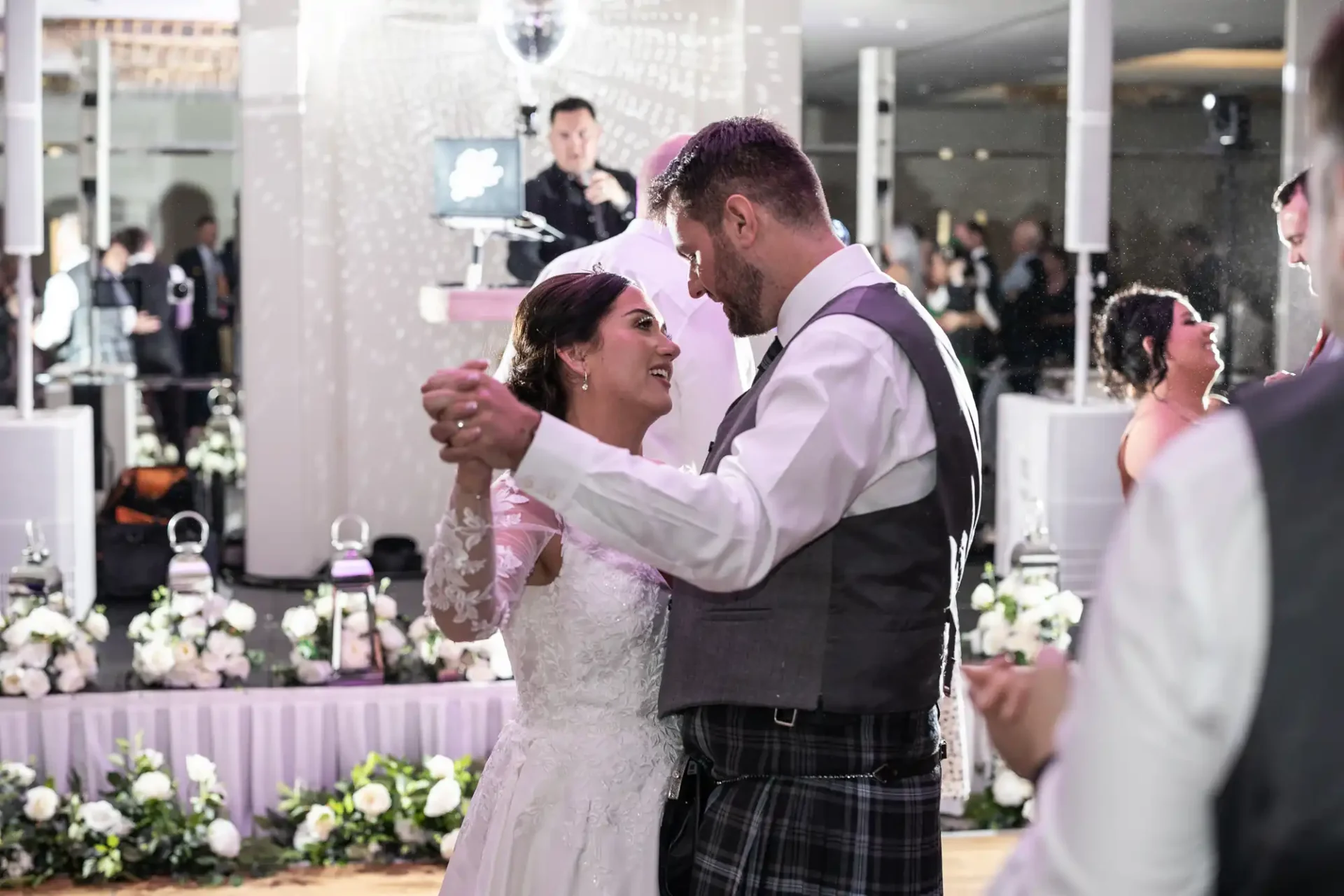 A bride and groom share a dance at their wedding reception, surrounded by floral decorations, with guests and a DJ in the background.