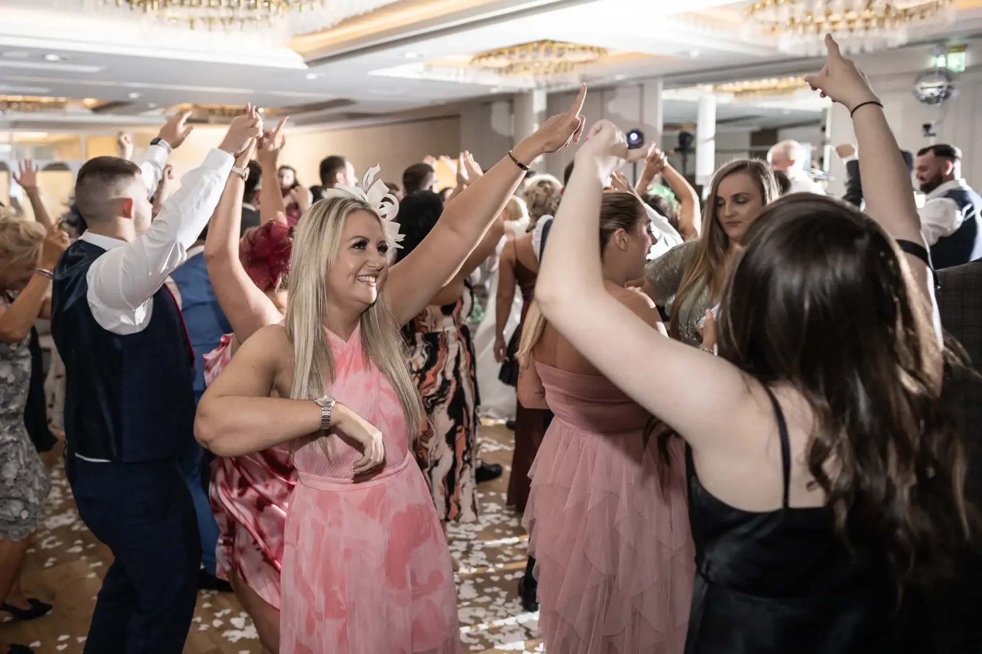 People dancing in a crowded indoor event space, some wearing formal attire. Confetti is scattered on the floor, and chandeliers hang from the ceiling.