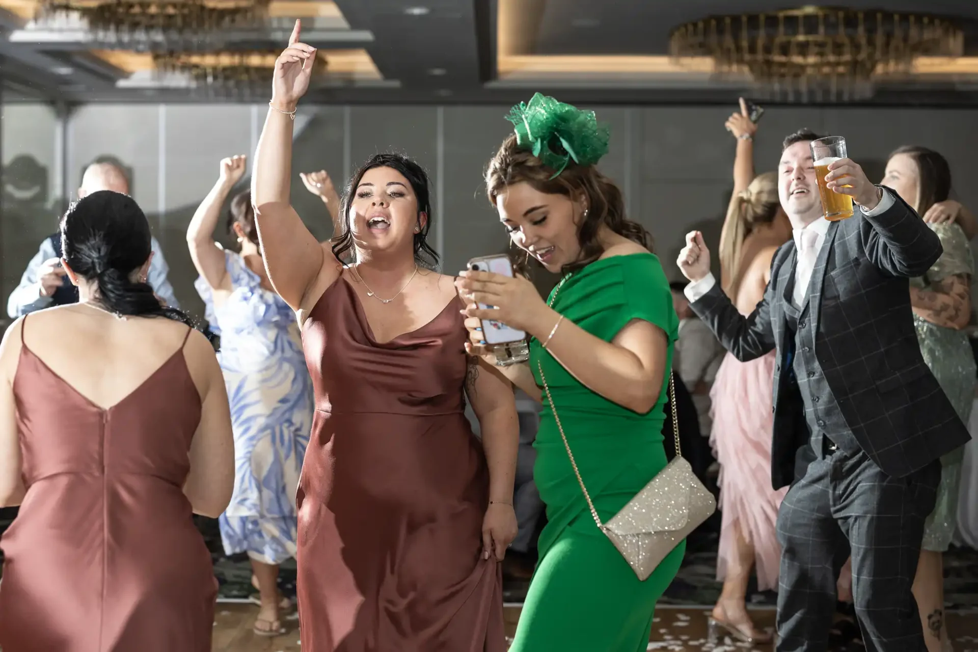 Guests dancing energetically at a celebration; a woman in a brown dress raises her hand, while another in a green dress checks her phone. Decorations and drinks are visible in the background.