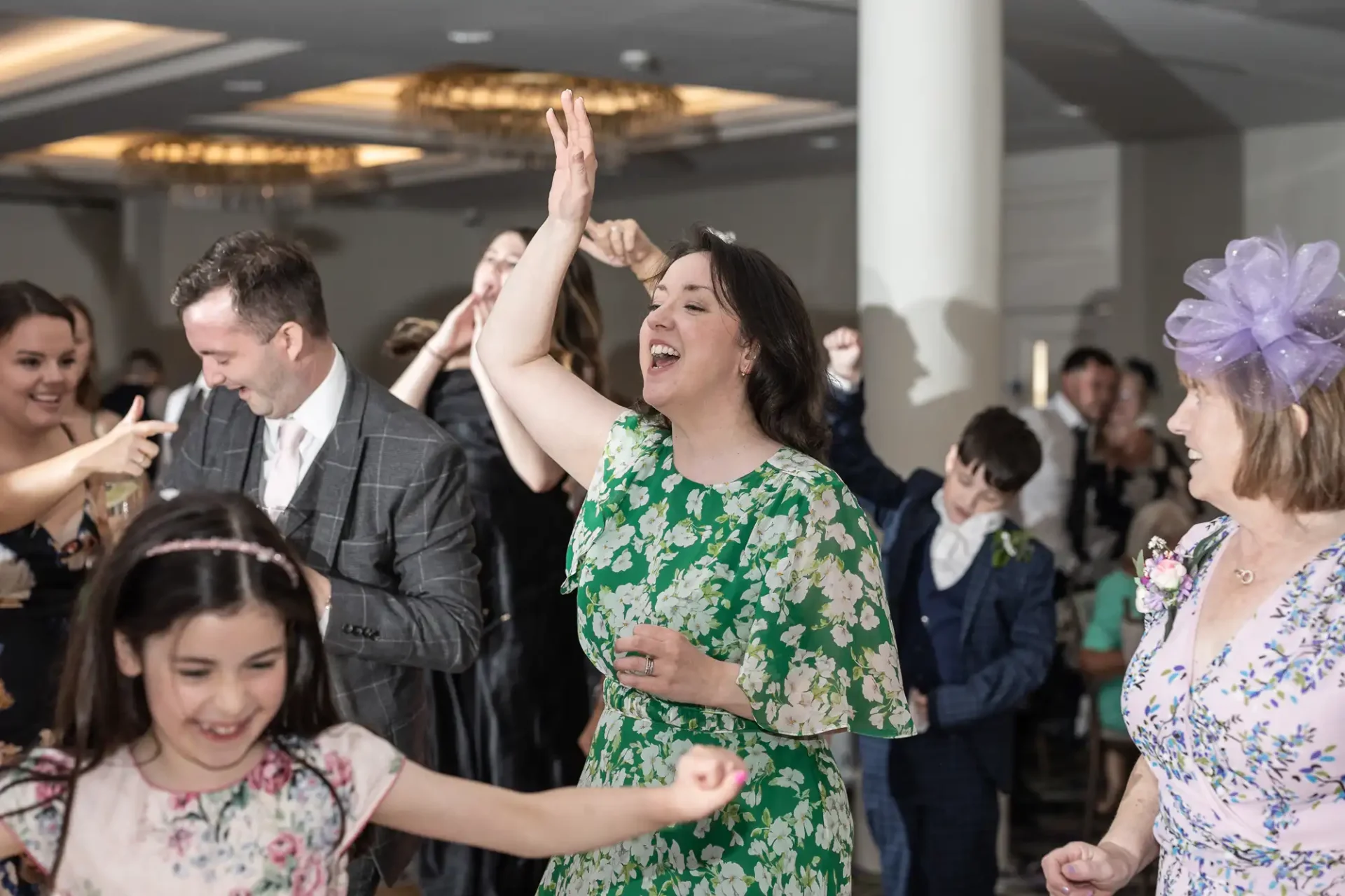 People dancing and celebrating at a lively event indoors, with a woman in a green floral dress prominently enjoying the moment.