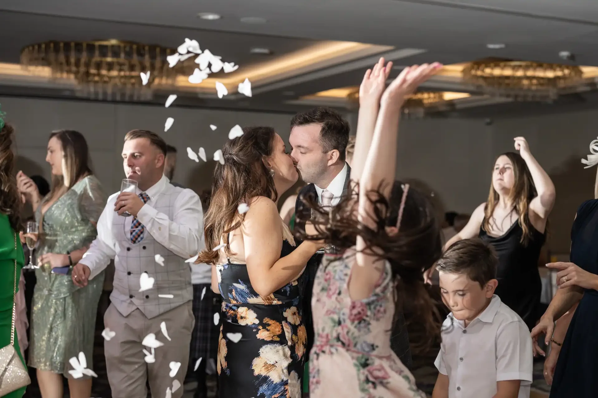 Couple kissing while people dance around them at a party; petals thrown in the air.