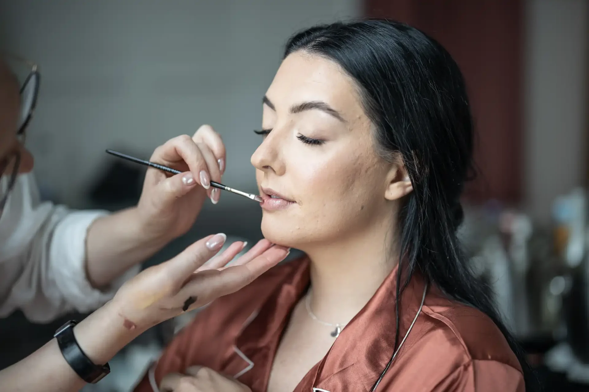 A woman with dark hair wearing a brown robe is having lipstick applied by a person with a brush. Her eyes are closed.