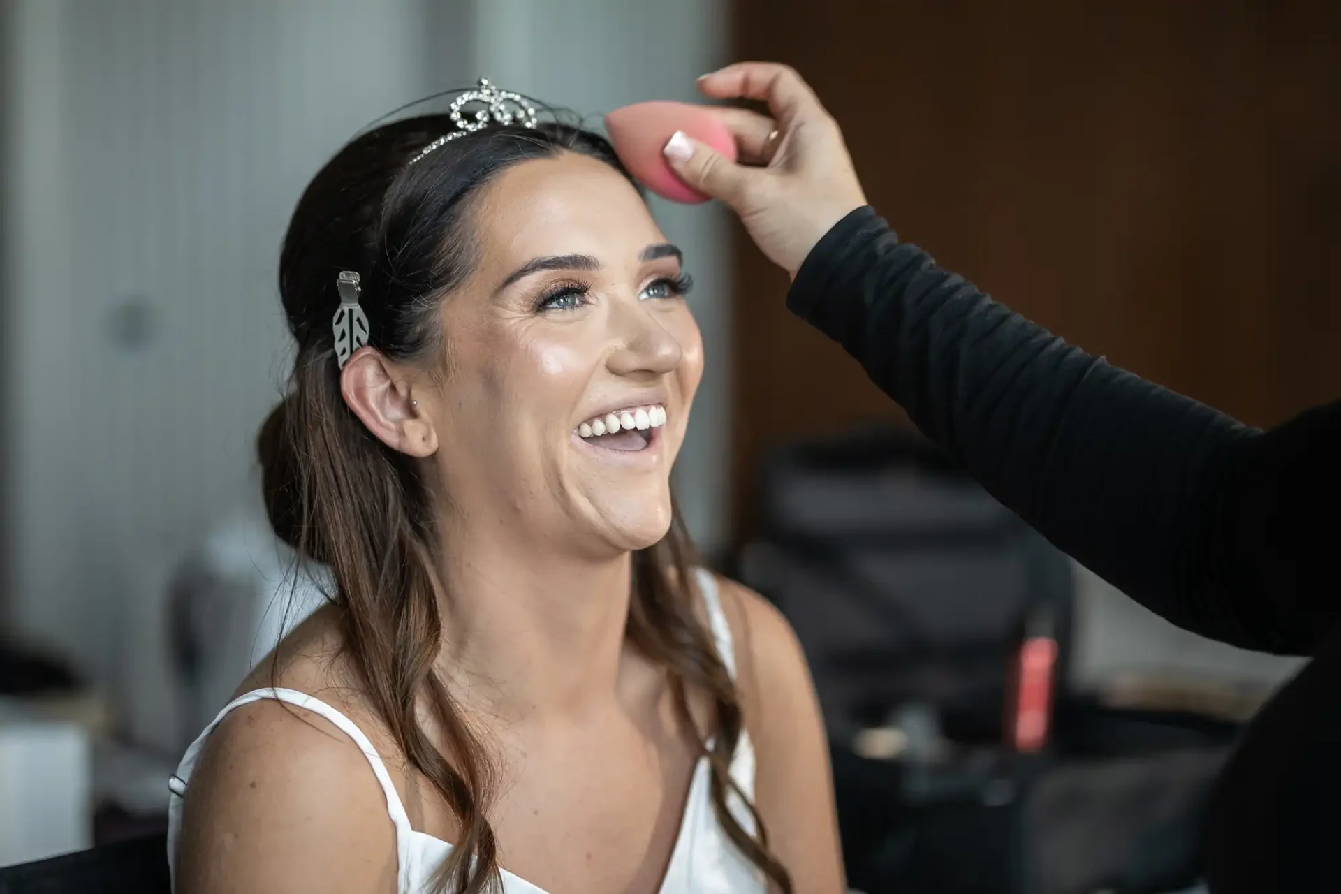 A woman in a white top smiles as a person uses a makeup sponge on her forehead. She wears a tiara and hairpins.