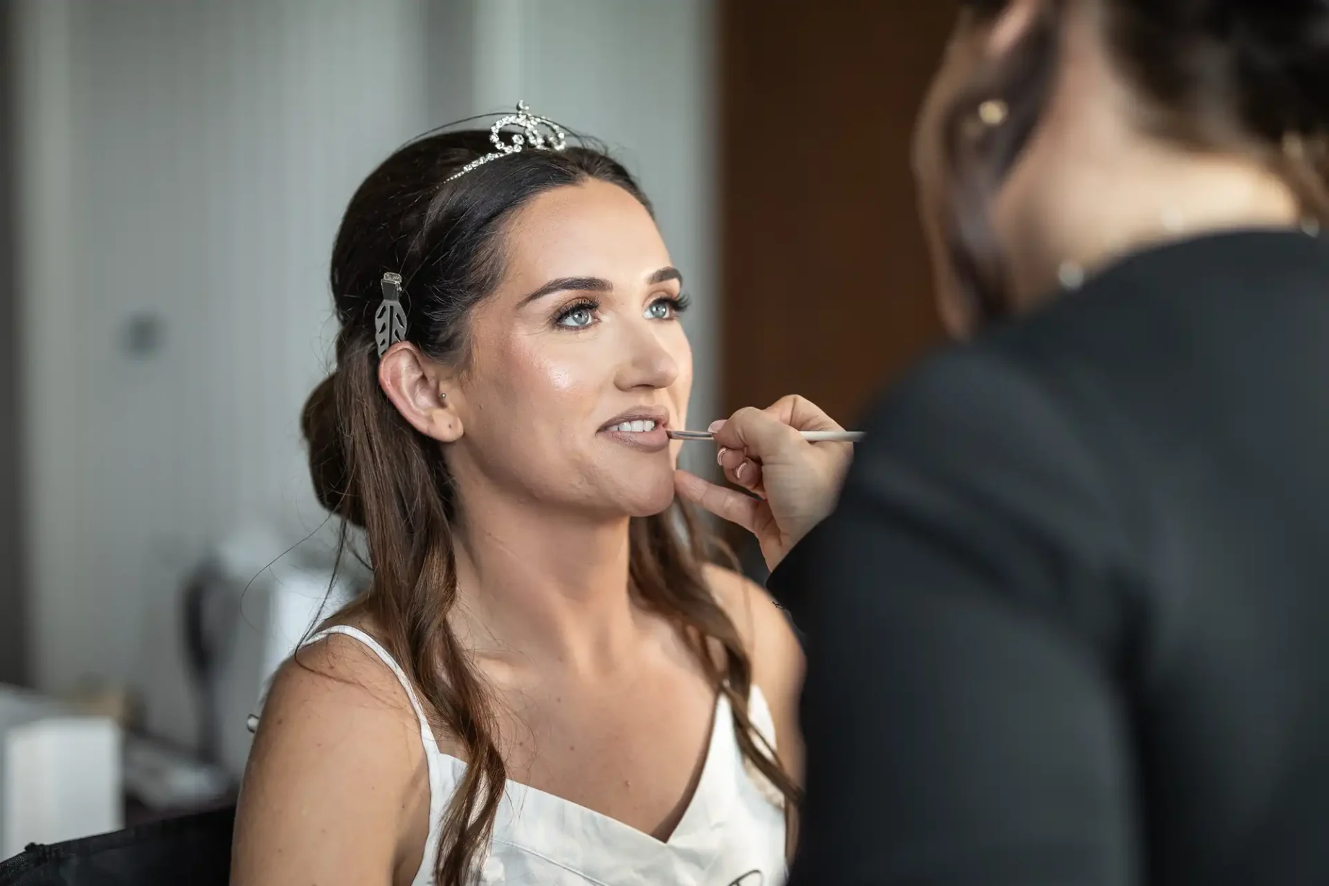 Woman having lipstick applied by a makeup artist, wearing a white dress and headpiece, seated indoors.