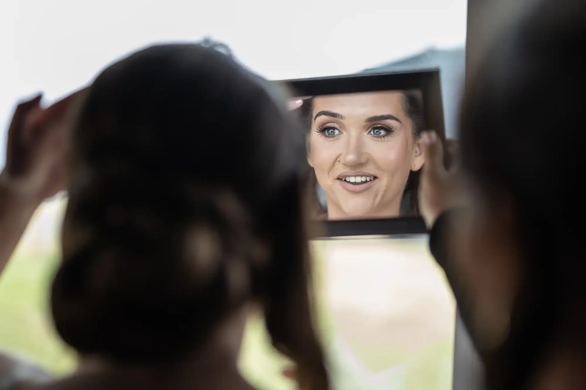 A woman holding a mirror reflects another woman with makeup on, smiling.