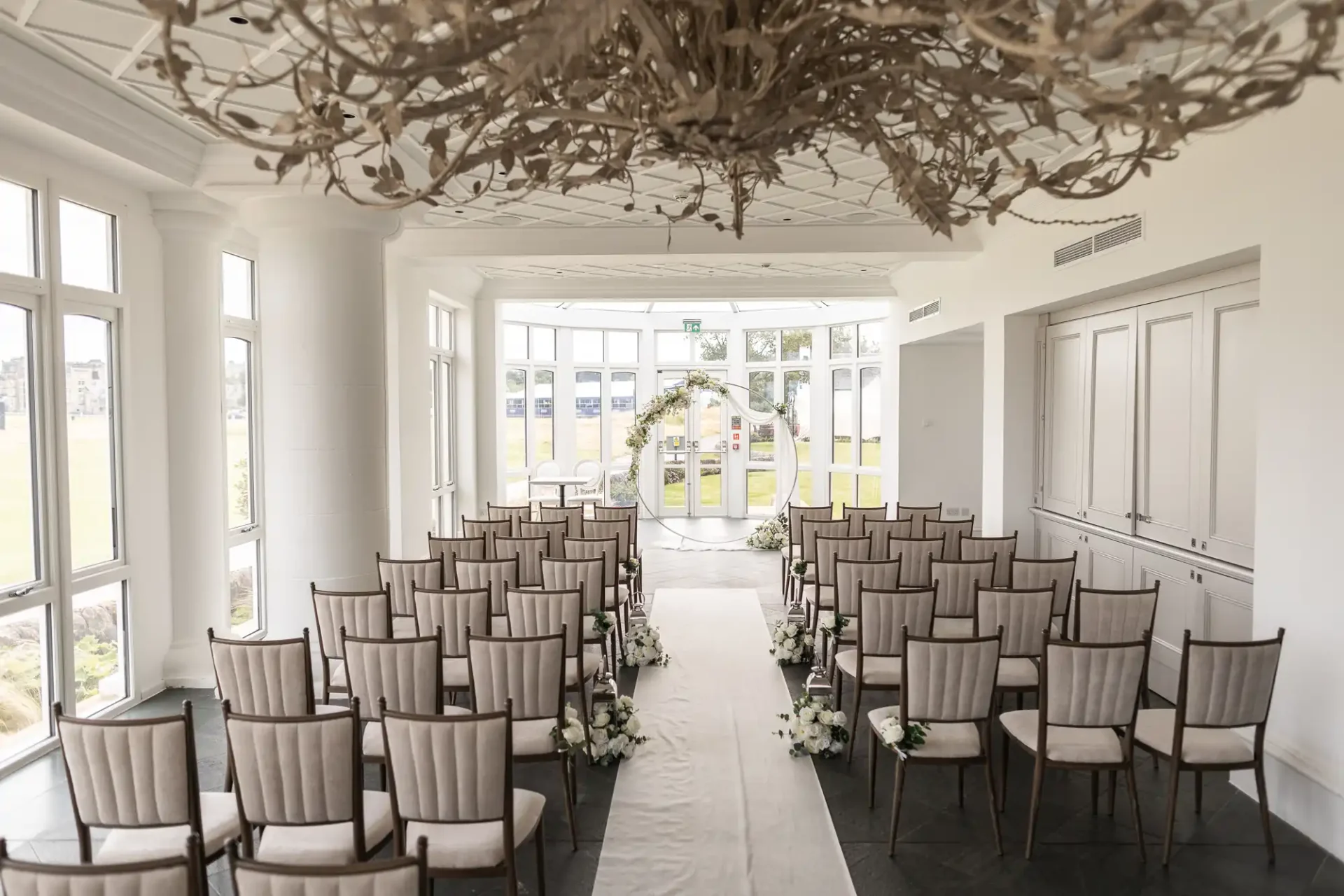 An indoor wedding venue with rows of chairs facing an arch decorated with flowers. Large windows line the room, allowing natural light to fill the space.