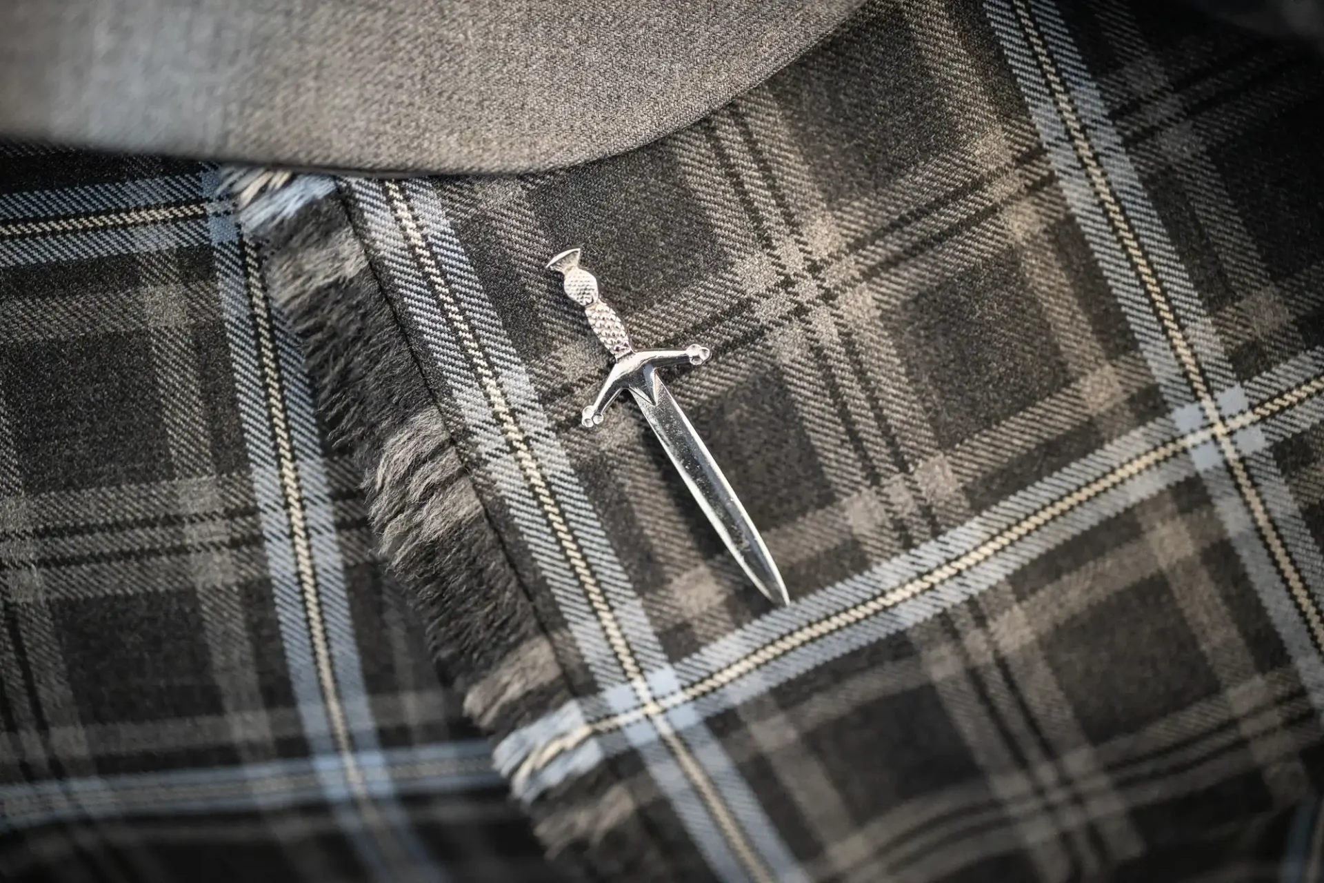 A silver sword-shaped brooch is pinned to a gray and black plaid fabric.