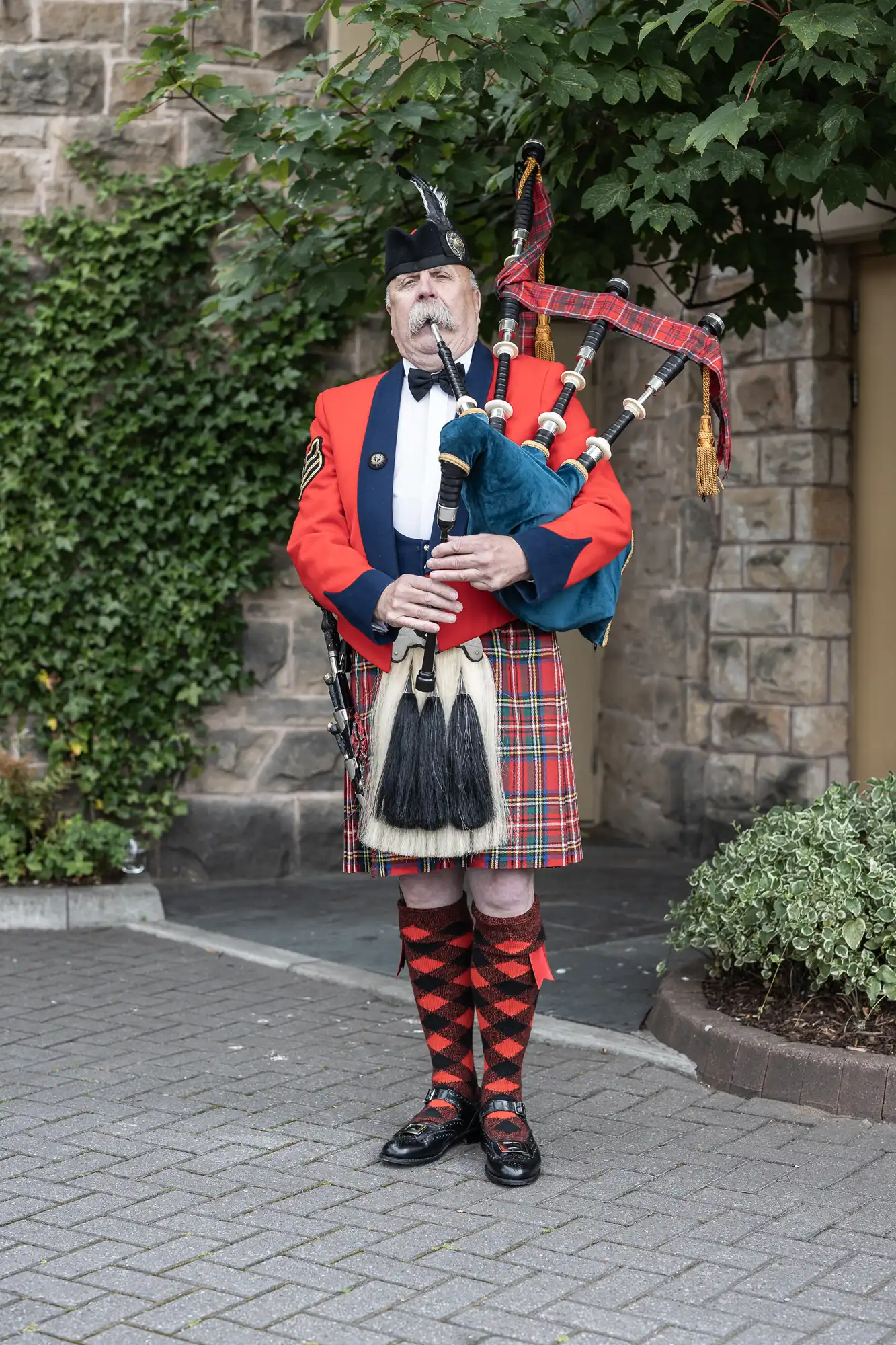 Man in traditional Scottish attire plays bagpipes outdoors, standing on a stone path by a vine-covered wall.